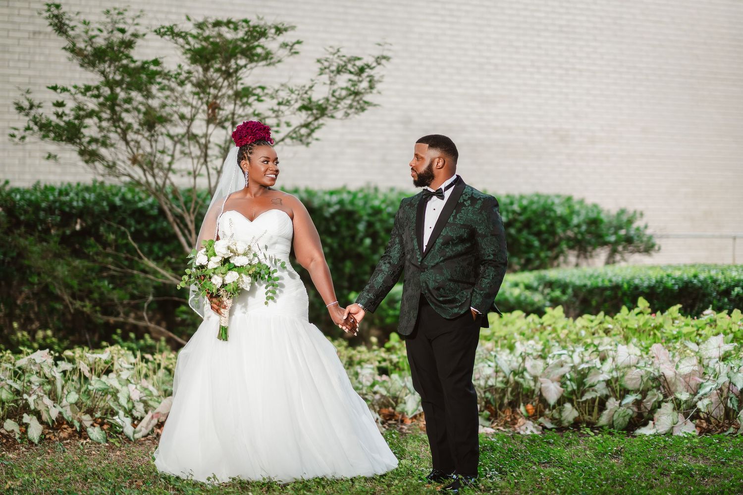 Wedding couple poses together outdoors against white wall with garden greenery.