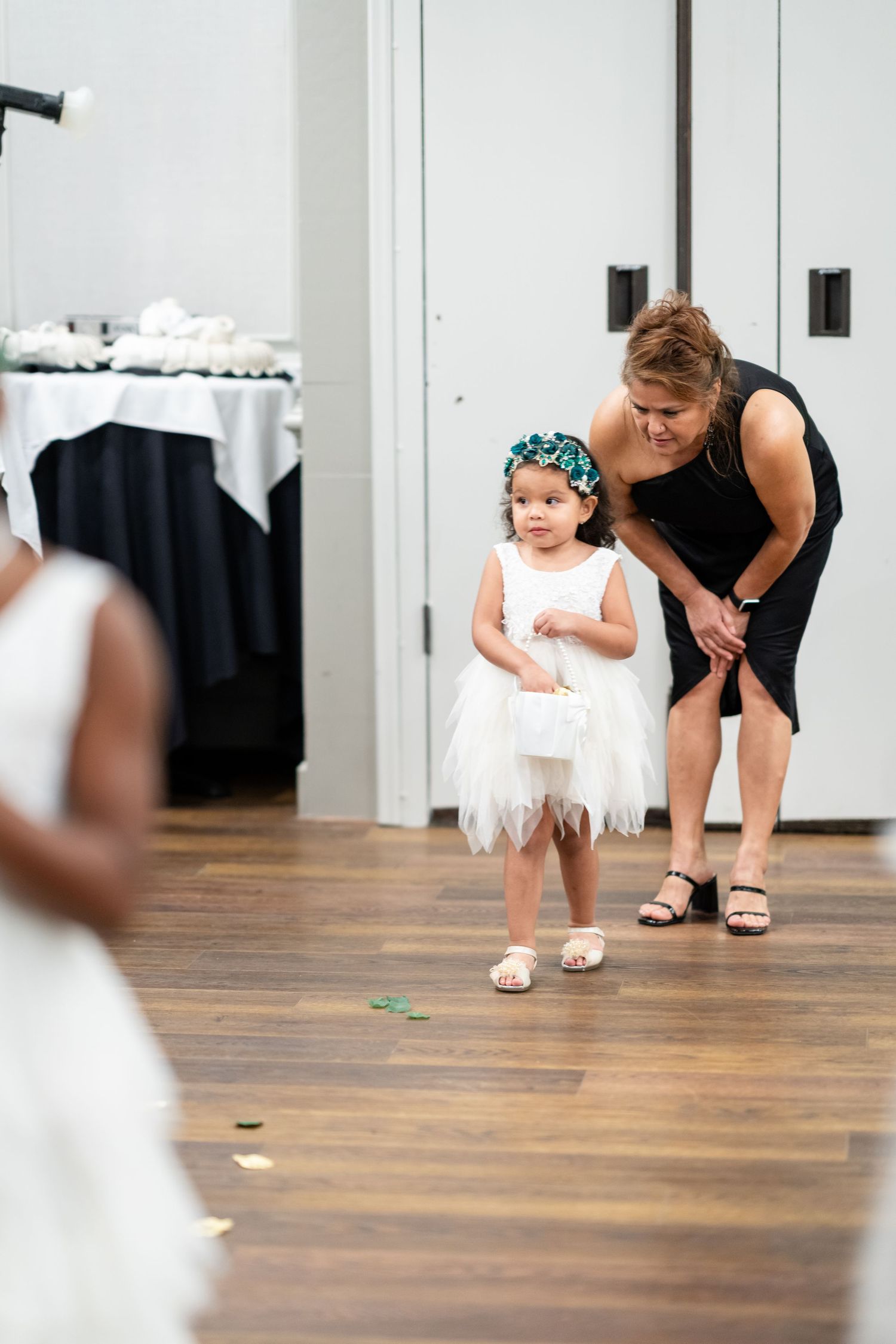 A young child in a white tulle dress and turquoise headband stands with an adult on a hardwood floor at an event.