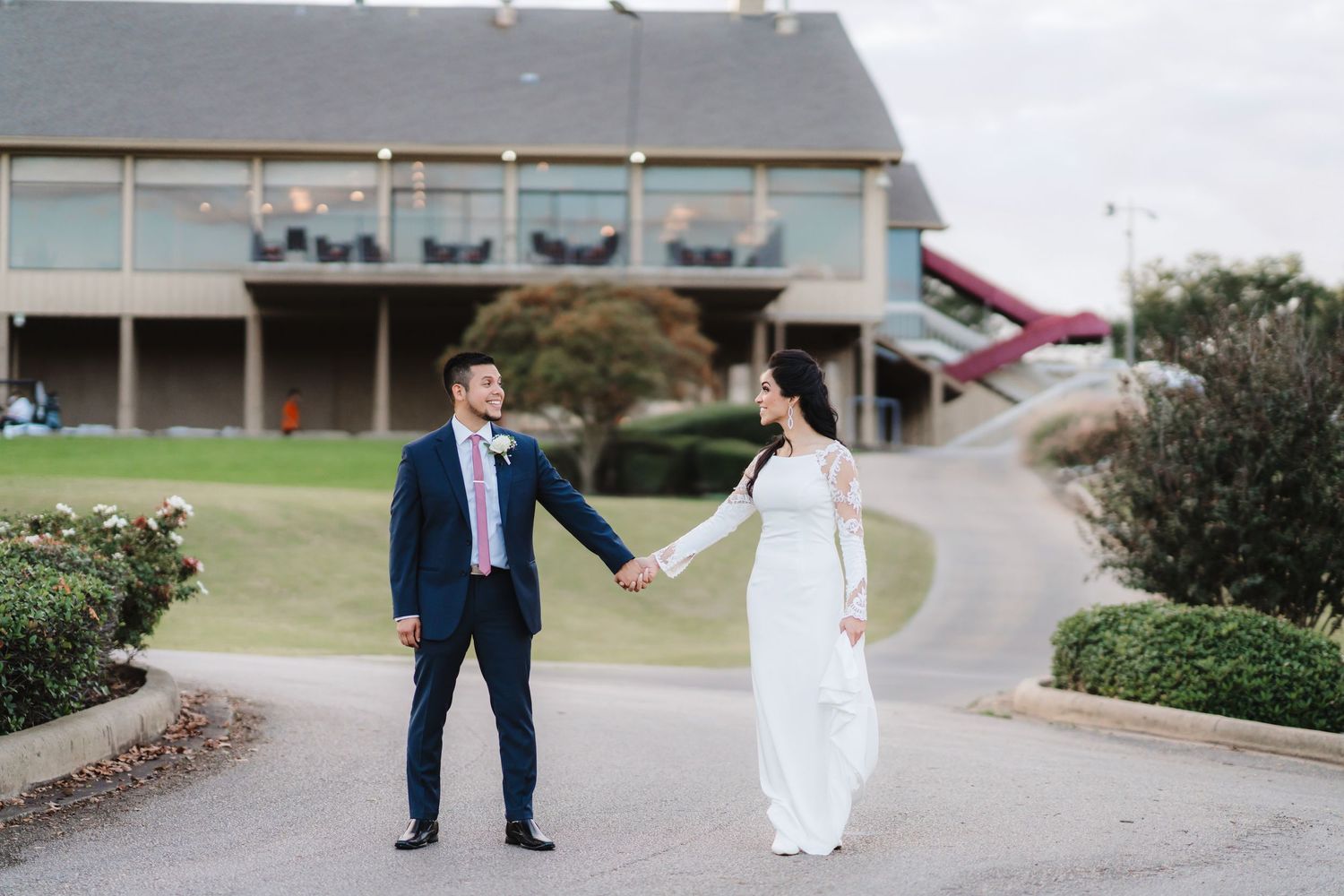 Wedding couple walks together outdoors on a path near modern buildings and landscaping.