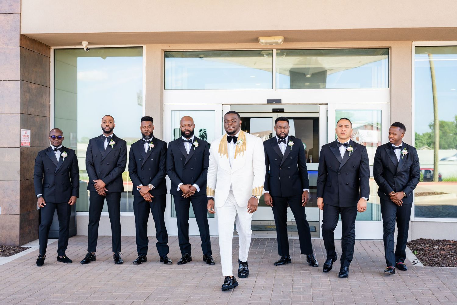 A wedding party in formal black tuxedos and white suits stands in a line against a beige building exterior.