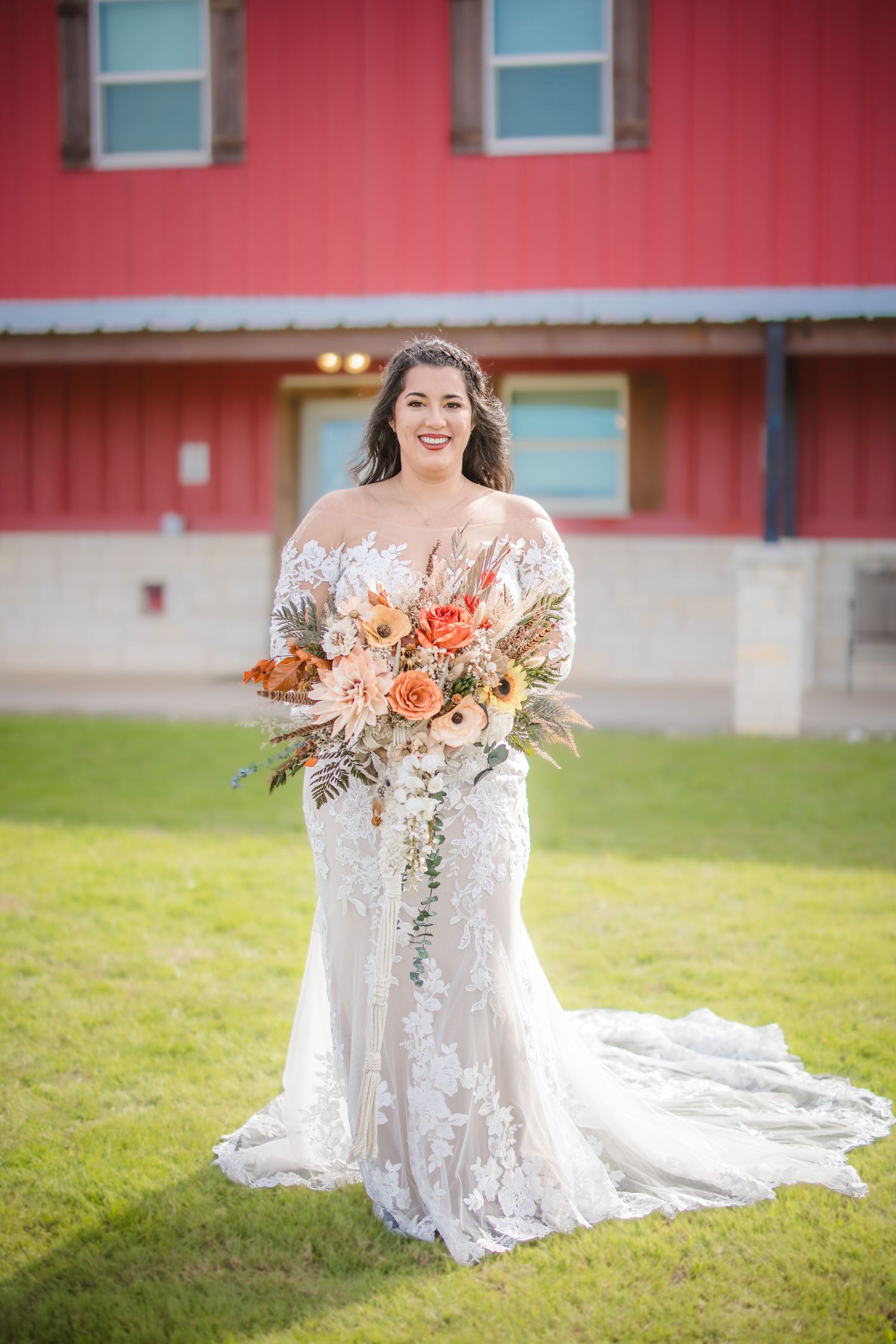 A bride in a lace wedding dress poses outdoors holding a coral and peach floral bouquet against a red building.