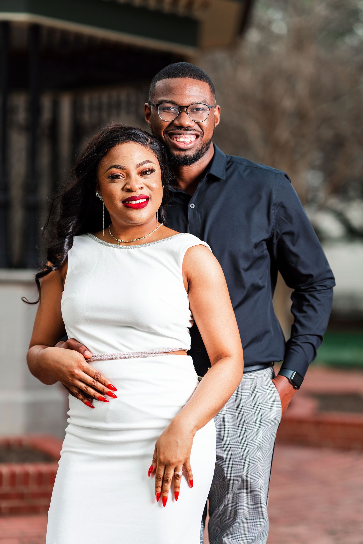 A couple poses together outdoors, with one person wearing a white sleeveless dress and the other in a dark button-down shirt.