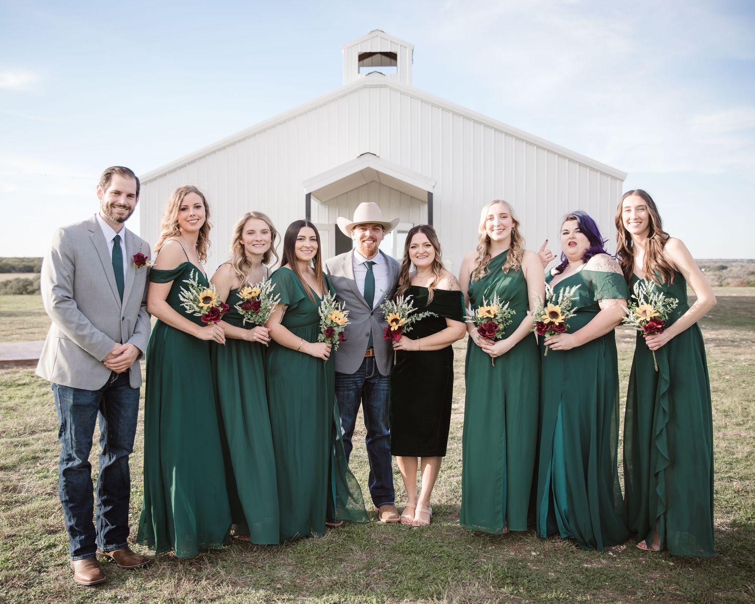 Wedding party in emerald green dresses and gray suits posing together outside a white barn.