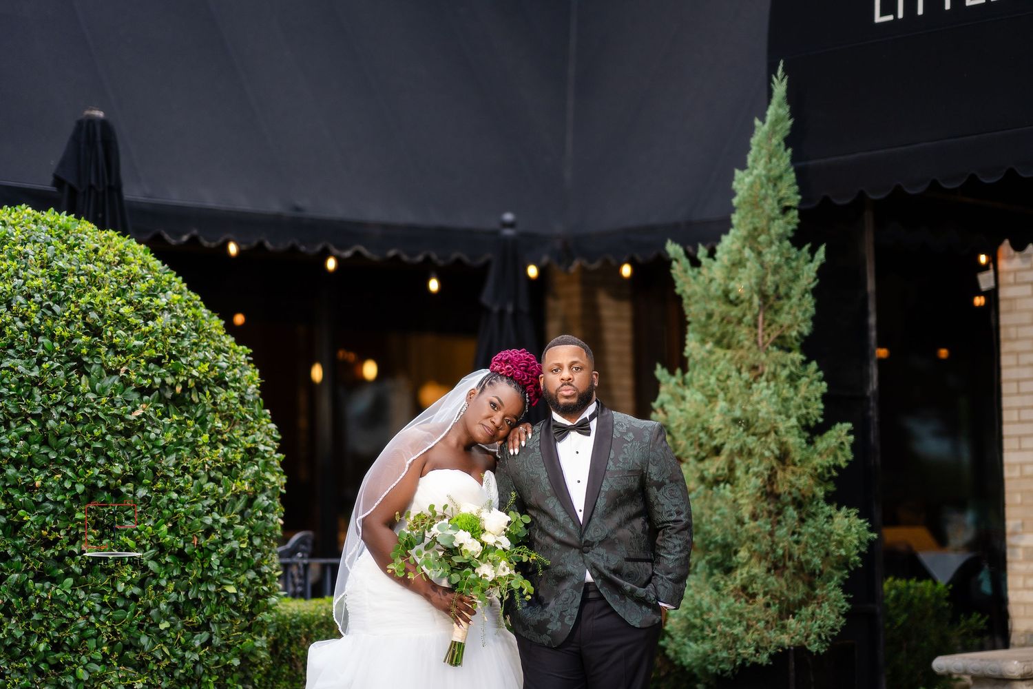 A wedding couple poses together outdoors against a backdrop of greenery and string lights.