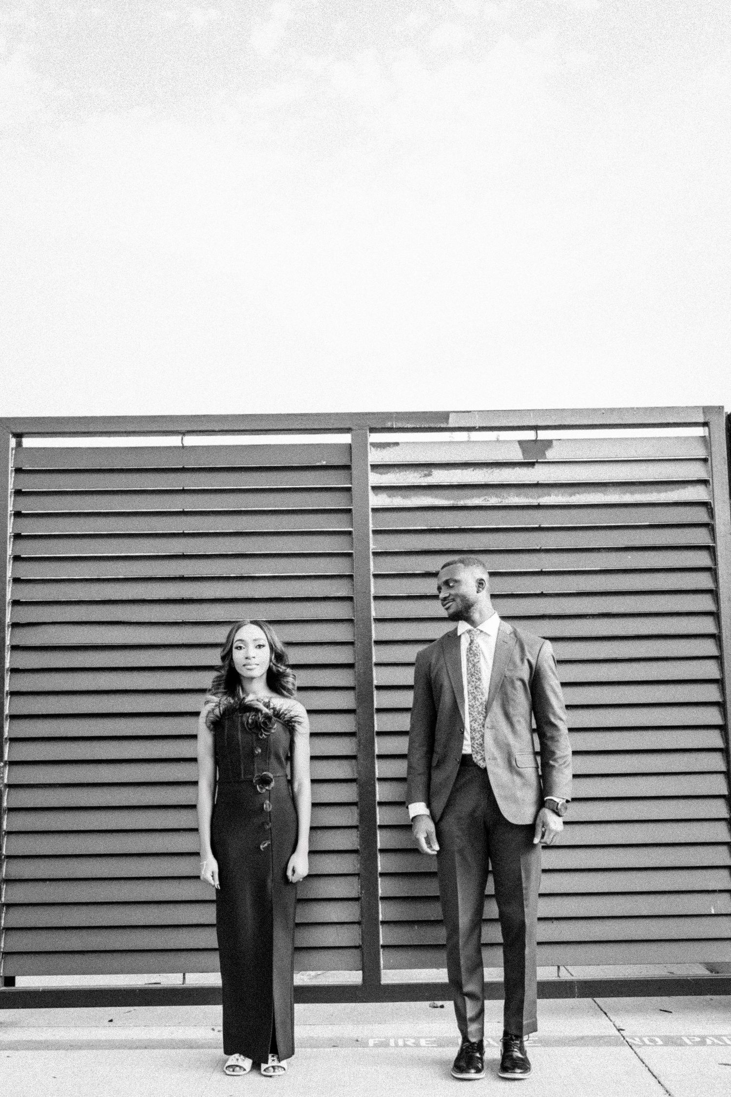 Black and white photo of a couple standing against a slatted wall in modern formal attire.