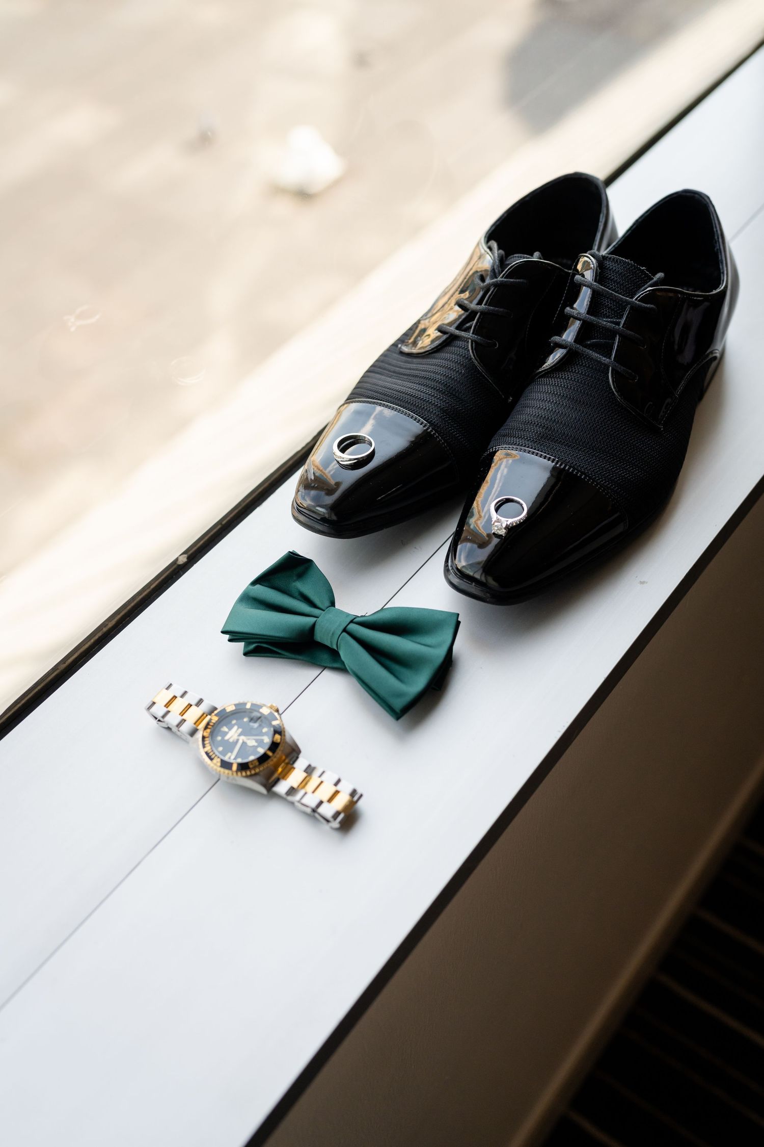 Wedding accessories including black dress shoes, green bow tie and watch on white surface.