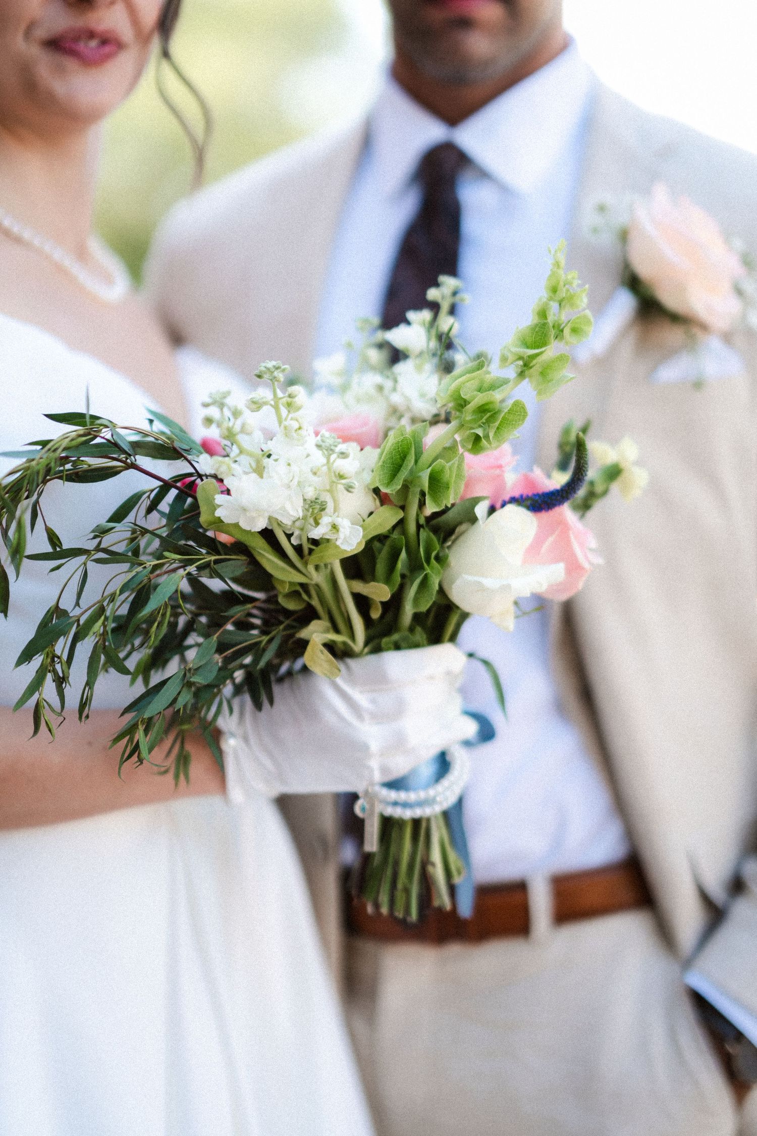 A wedding bouquet with white and pink flowers tied with ribbon shown against a light gray suit and white dress.