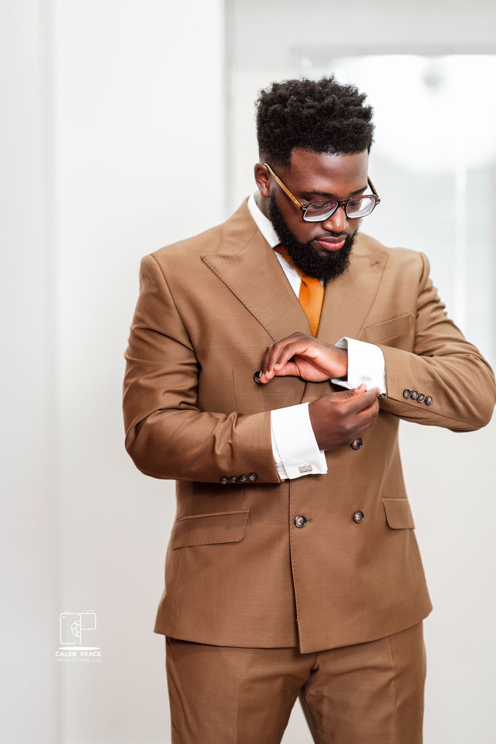 Professional in a tailored camel double-breasted suit adjusts cufflinks while wearing glasses and a neat beard.