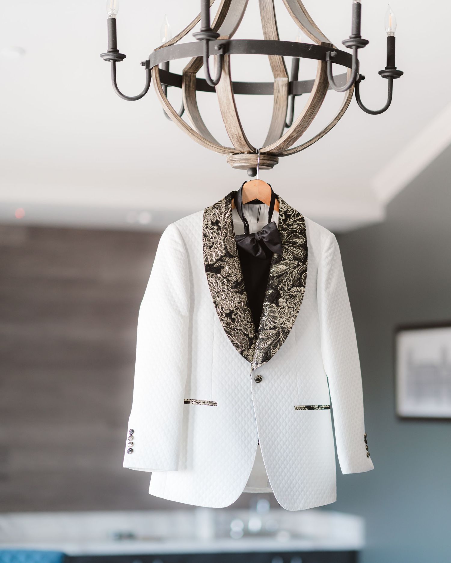 An elegant white tuxedo jacket with floral lapels hangs under a rustic chandelier.