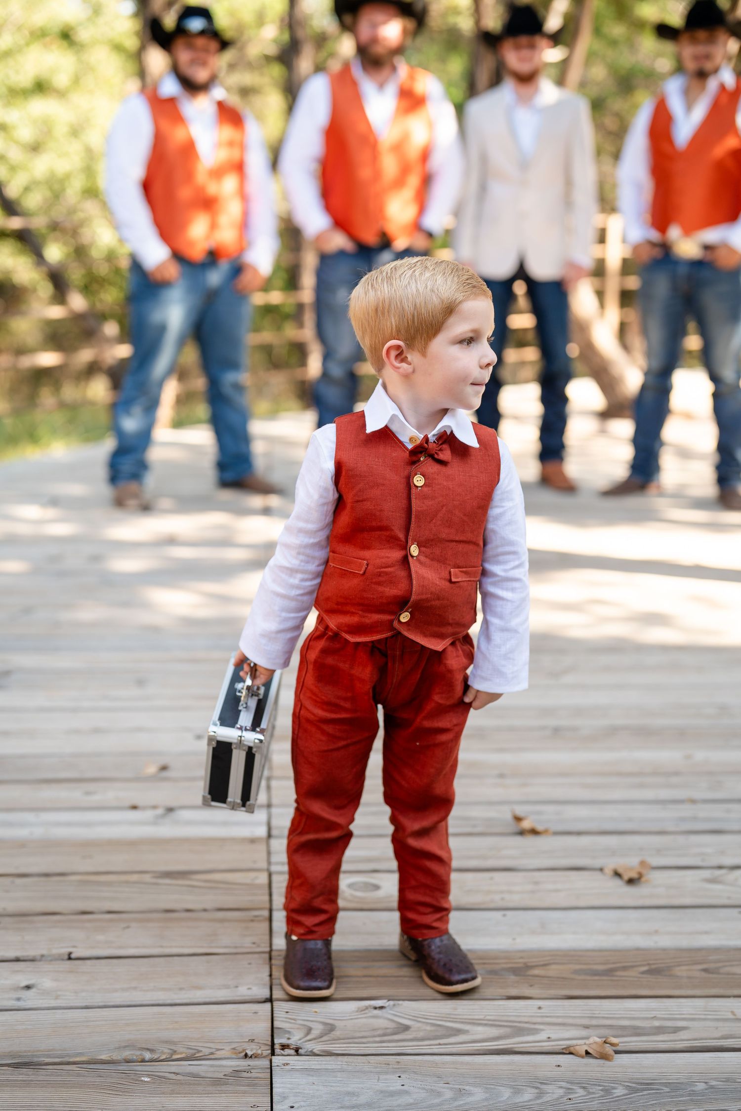 A small child in a red vest and white shirt walks on a wooden deck surrounded by adults in western wear.
