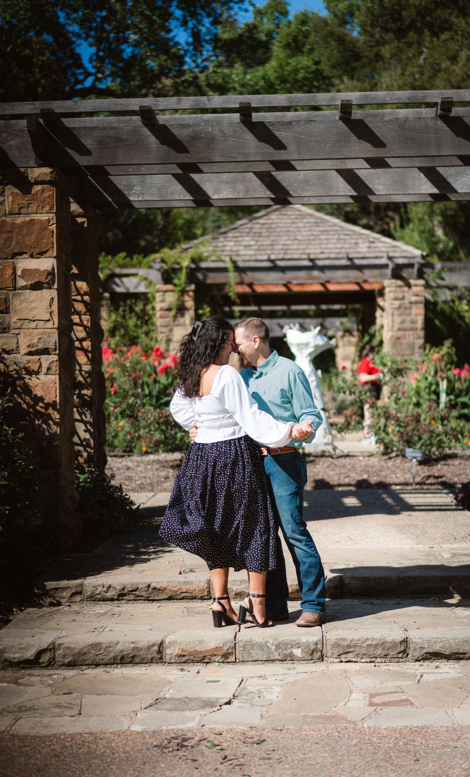 A couple sits together on stone steps in a garden setting under a wooden pergola on a sunny day.