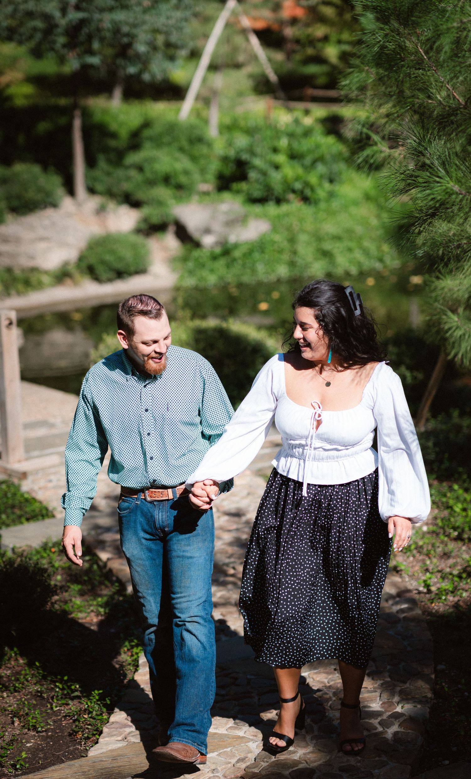 A couple walking hand in hand along a garden path surrounded by lush greenery on a sunny day.