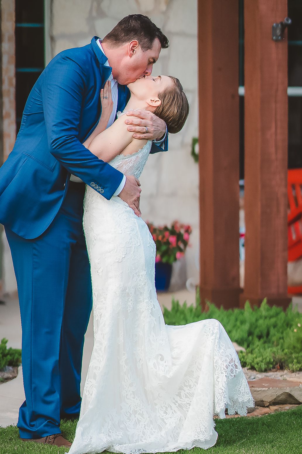 A romantic wedding moment captured outdoors with the couple in a blue suit and white lace gown sharing a kiss.