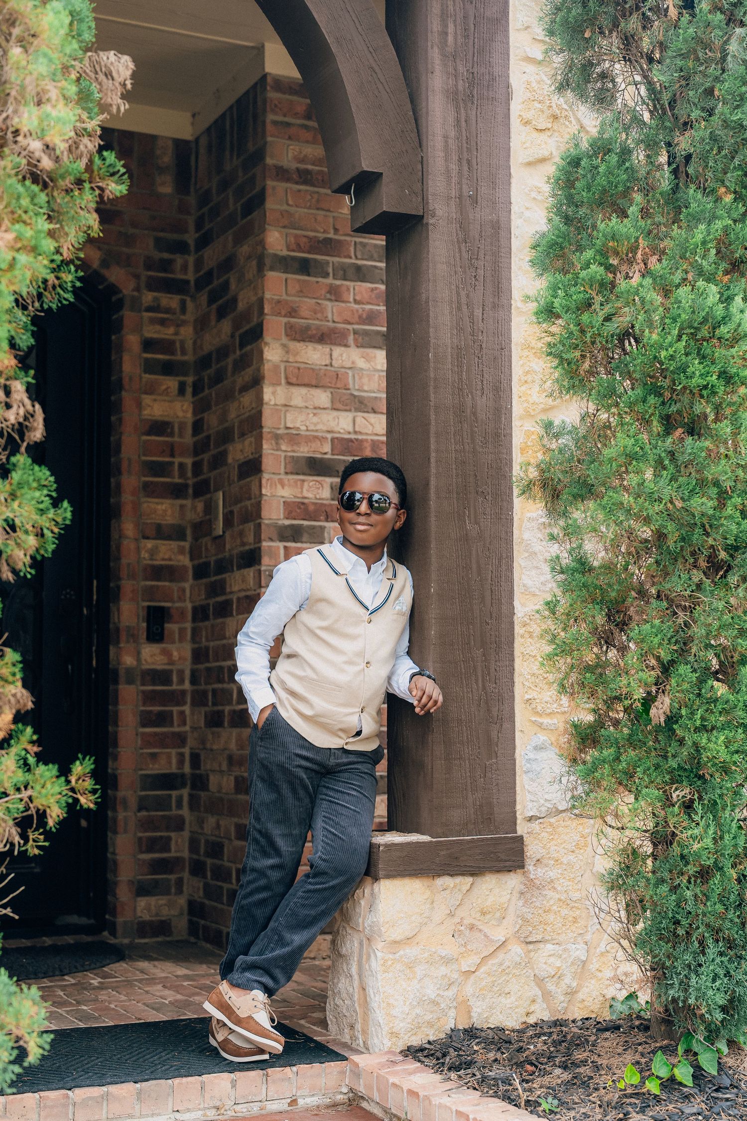 A person in a beige vest and sunglasses stands under an archway near brick columns and greenery.