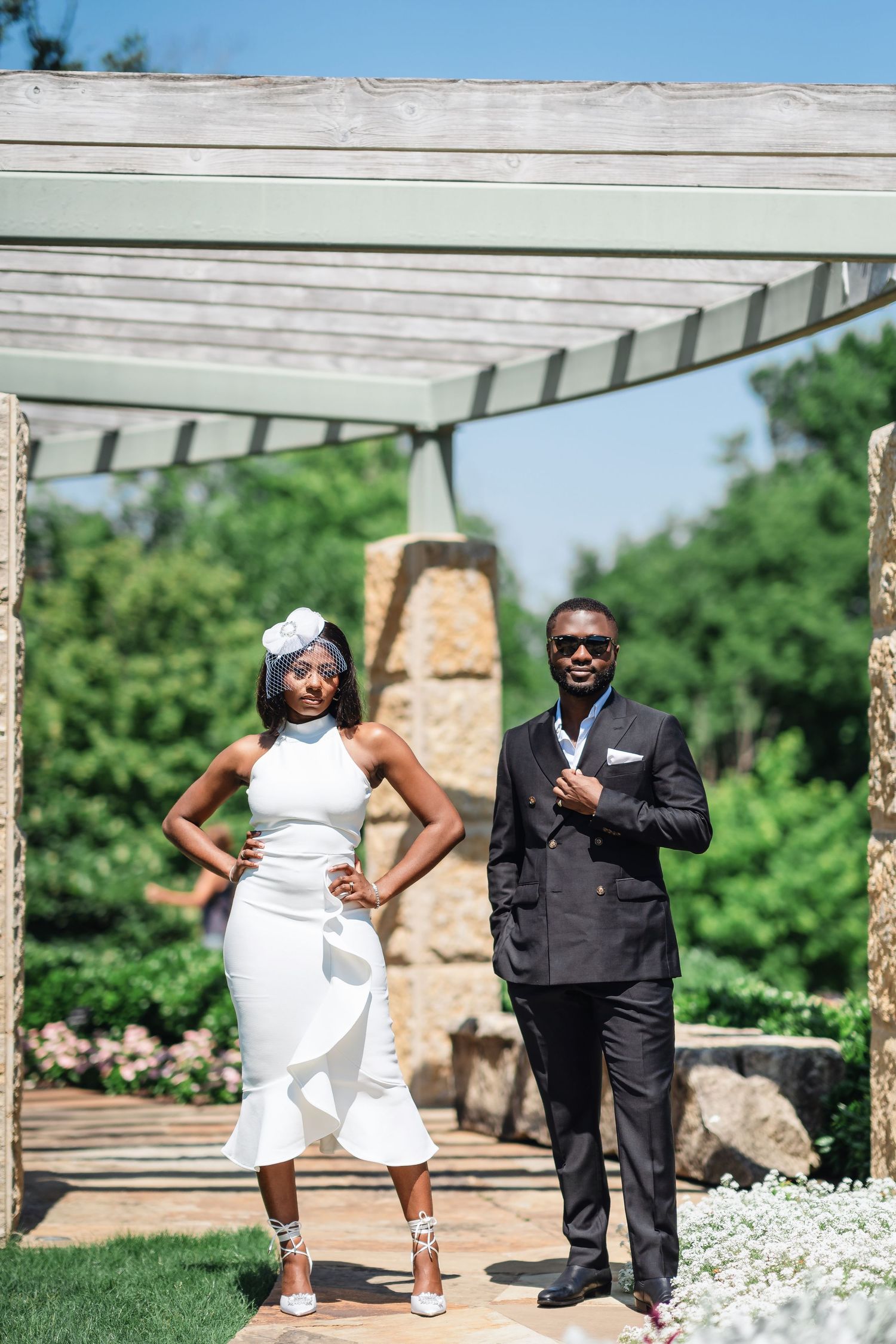 A stylish couple poses under a pergola in a garden setting wearing elegant wedding attire.