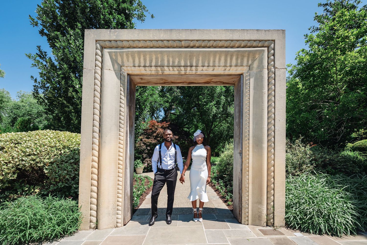 Wedding couple poses in an elegant outdoor archway with decorative stone frame surrounded by greenery.