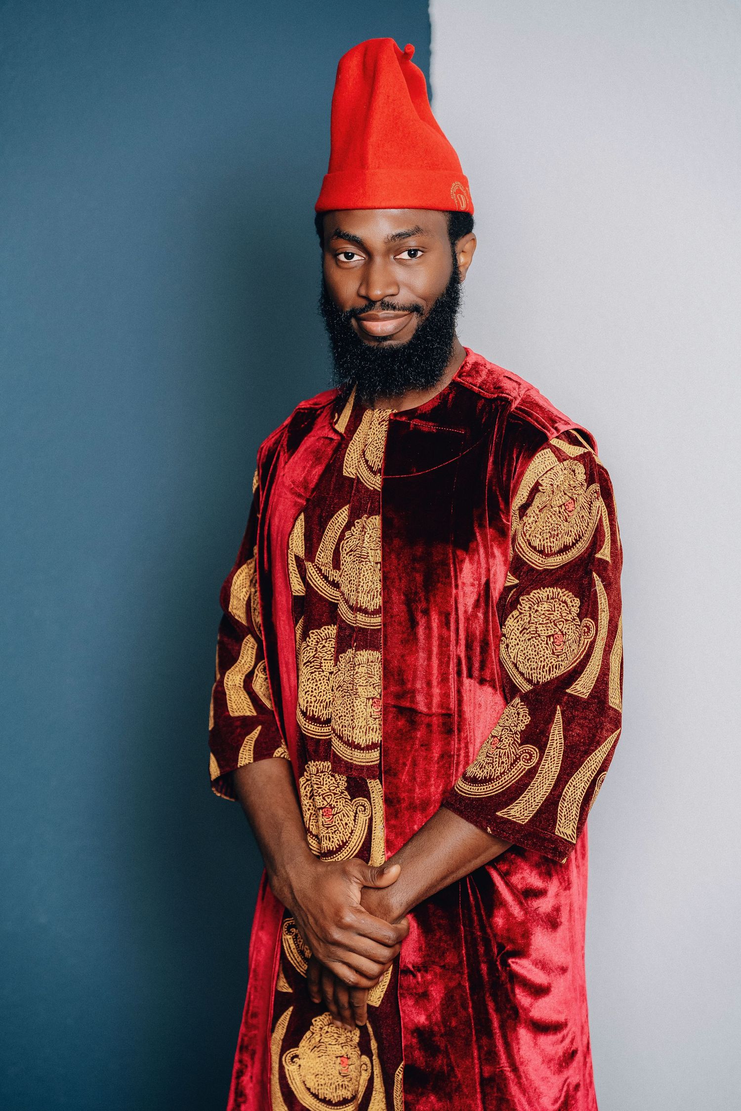 Traditional Nigerian attire featuring a red velvet robe with gold circular patterns and matching red cap against blue backdrop.