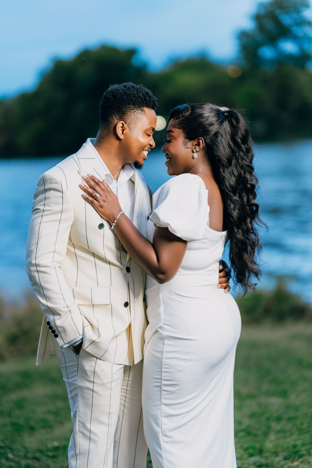 Couple in matching cream colored outfits embrace during lakeside photoshoot.