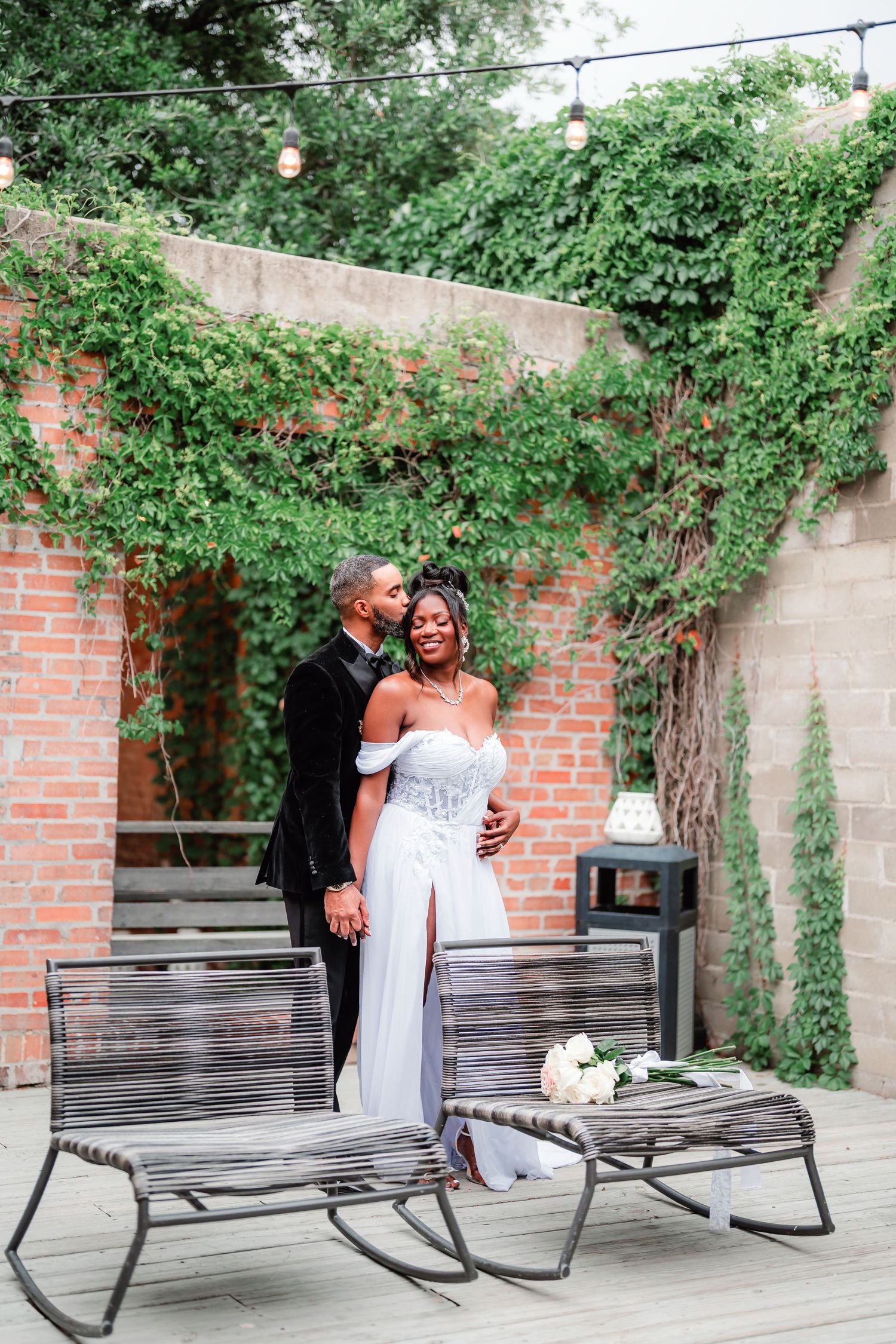 Wedding couple shares intimate moments on wooden benches against ivy-covered brick wall.