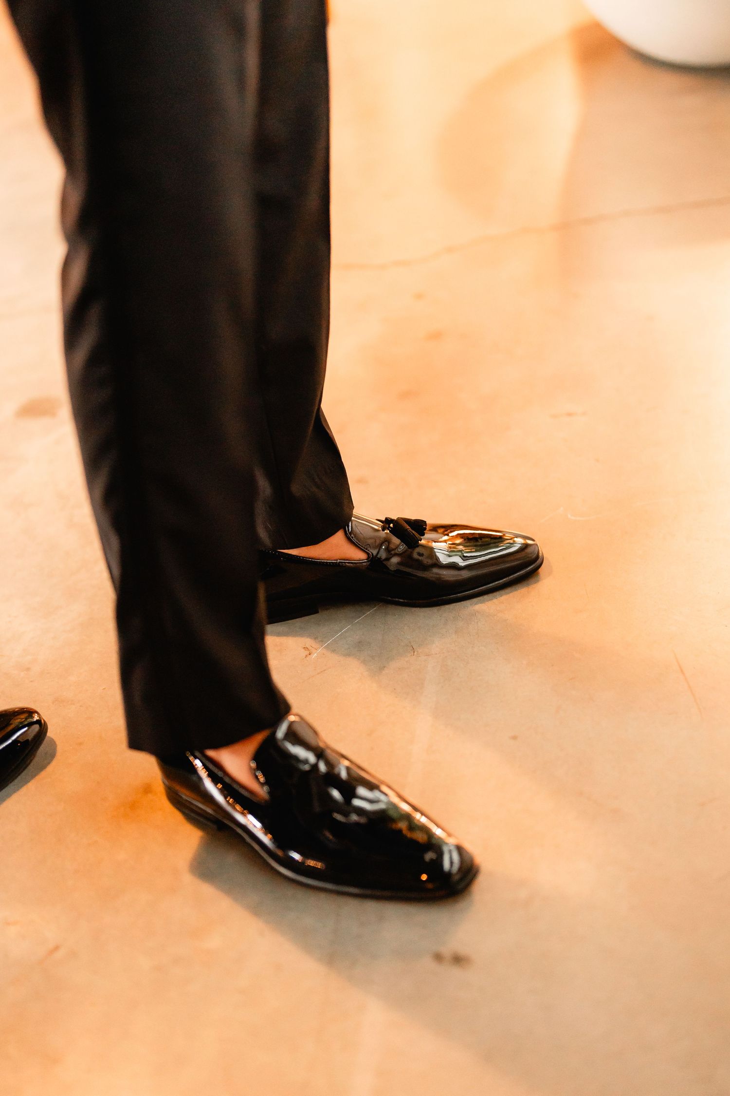 Close-up of shiny black formal dress shoes and pants on a beige floor.