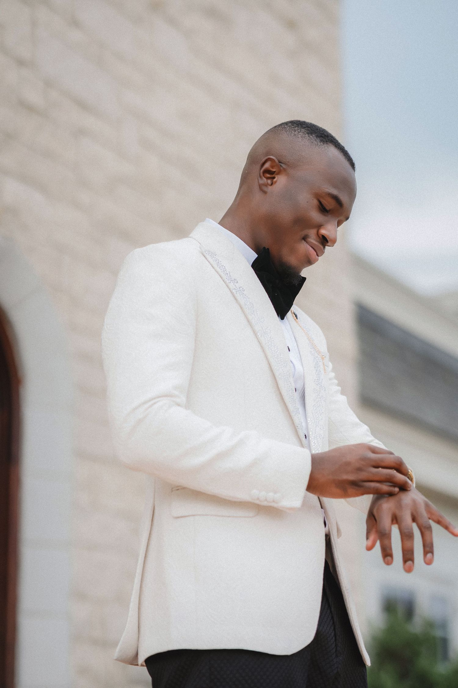 A groom in a white tuxedo jacket adjusts his cufflinks outdoors.