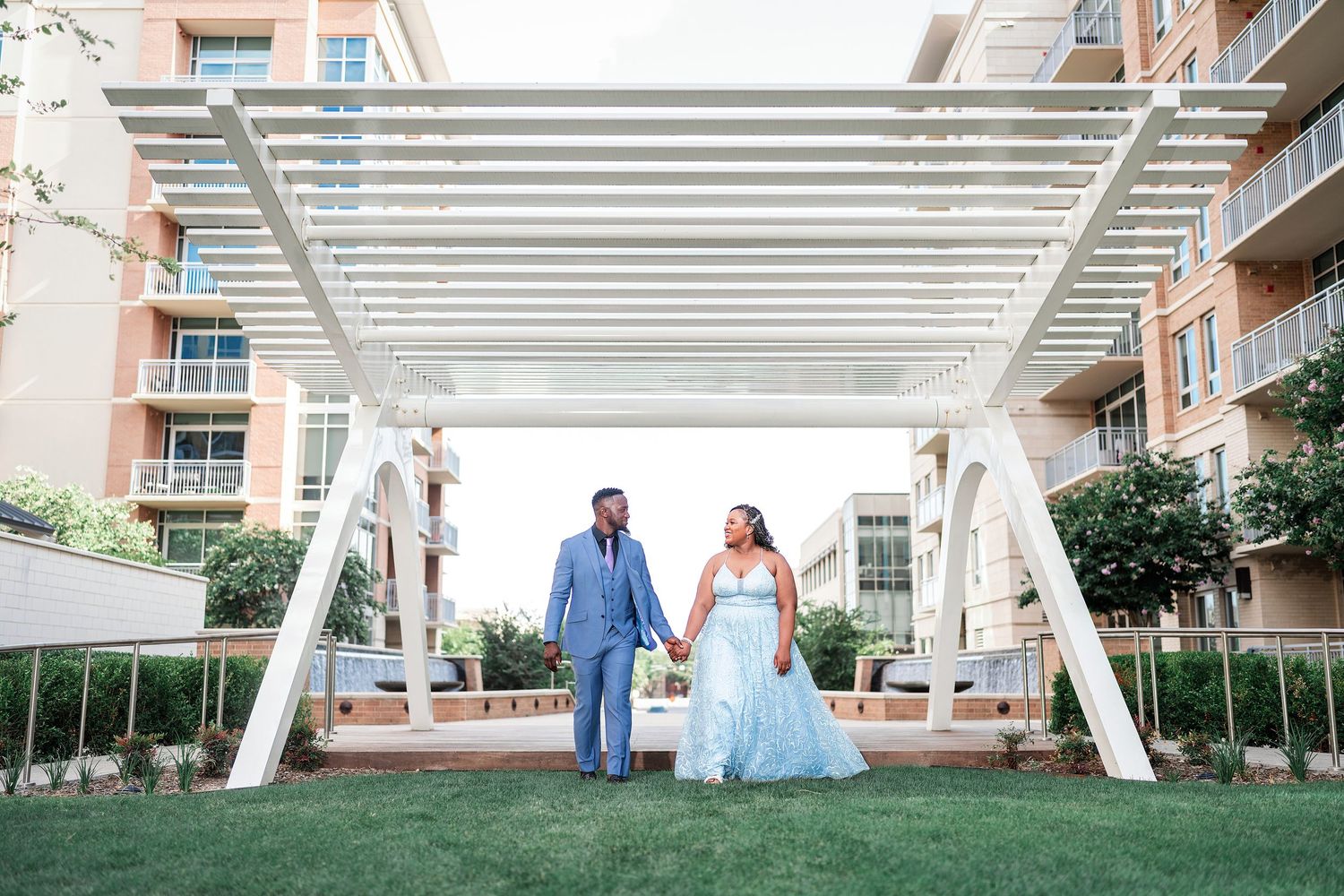 Couple walks together through a garden path surrounded by lush greenery and brick buildings on a sunny day.
