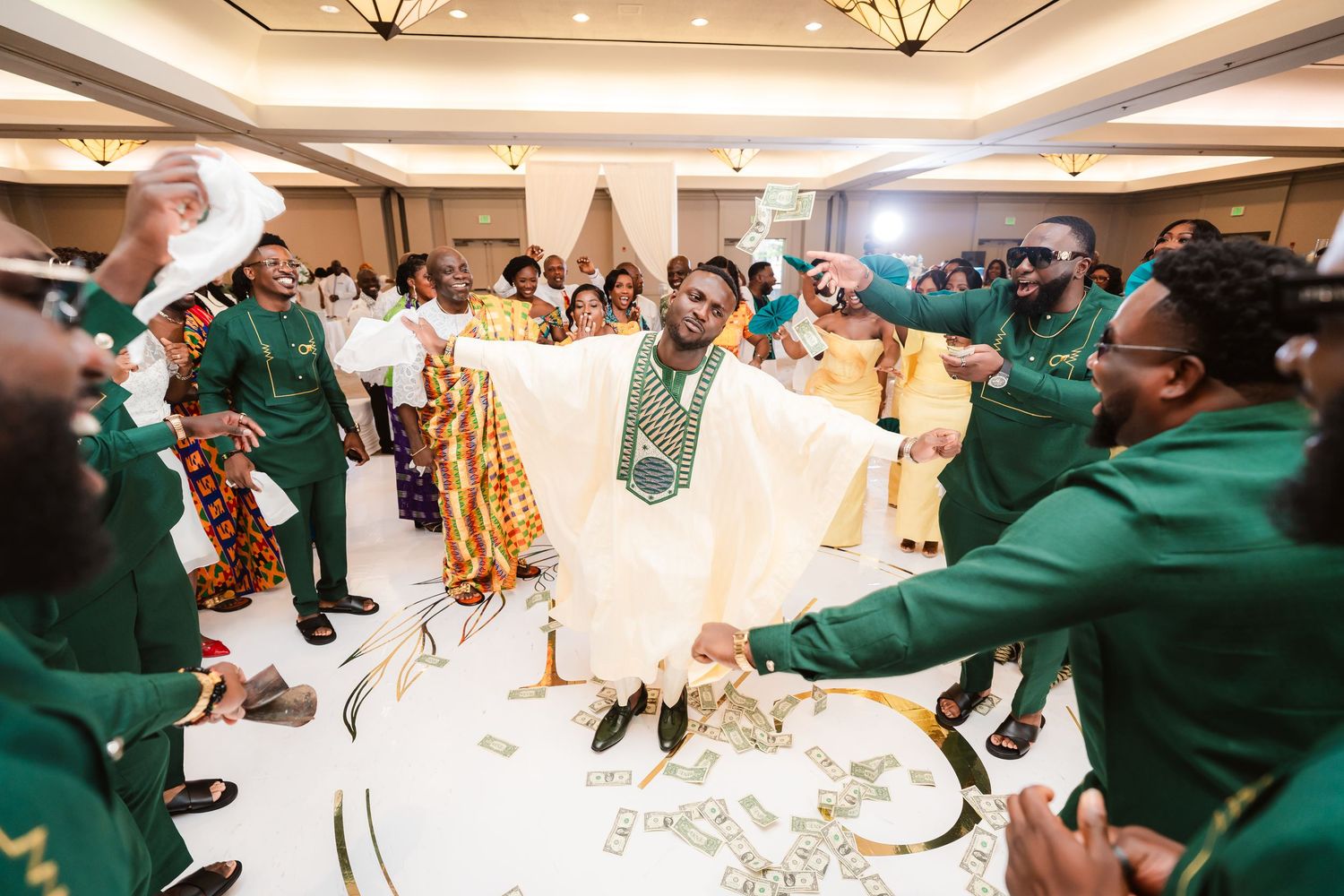 Wedding celebration with groomsmen in green suits dancing around the groom in white traditional attire.