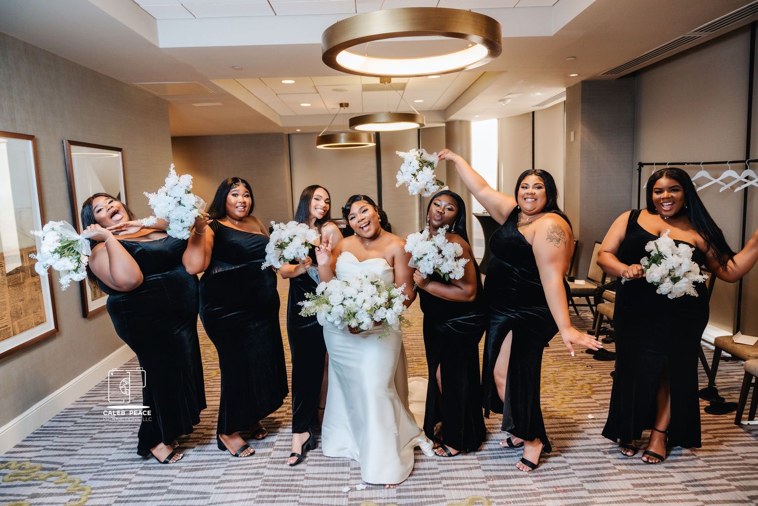 Bridesmaids in elegant black dresses pose with bride in white gown holding blue and white bouquets.