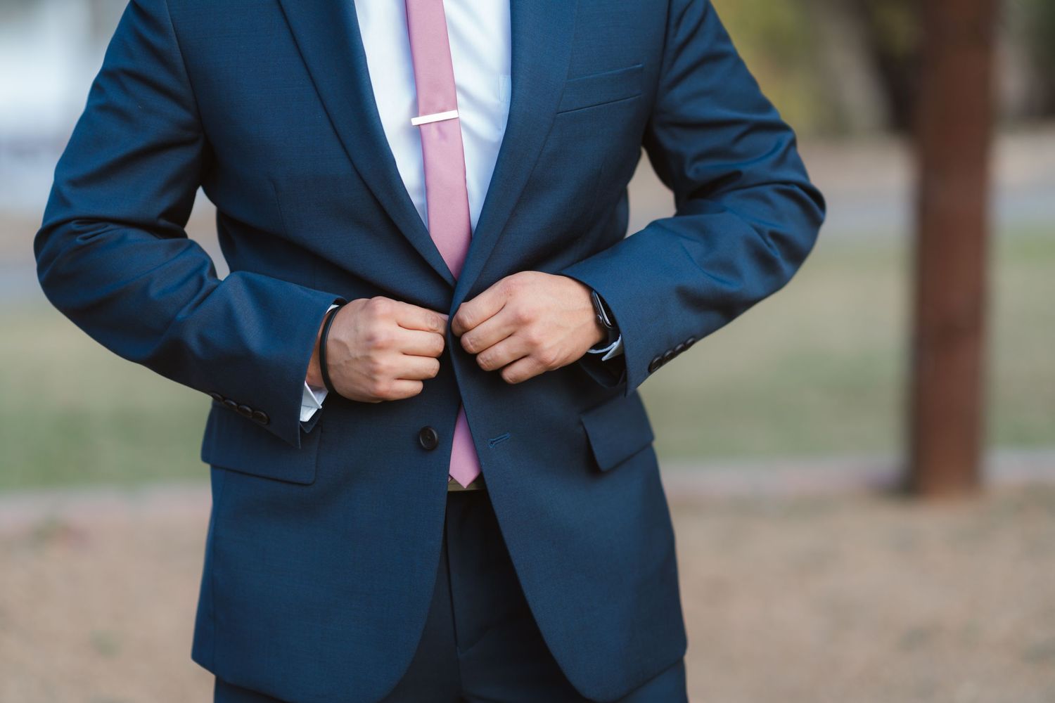 A person wearing a navy blue suit with a pink tie adjusts their jacket while standing outdoors.