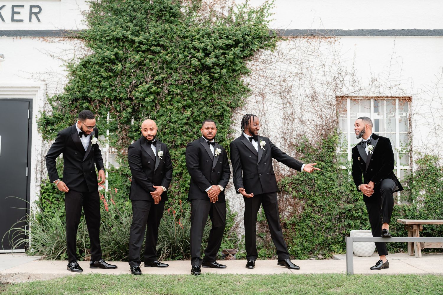 Groomsmen in black suits pose in sequence along a white building exterior.