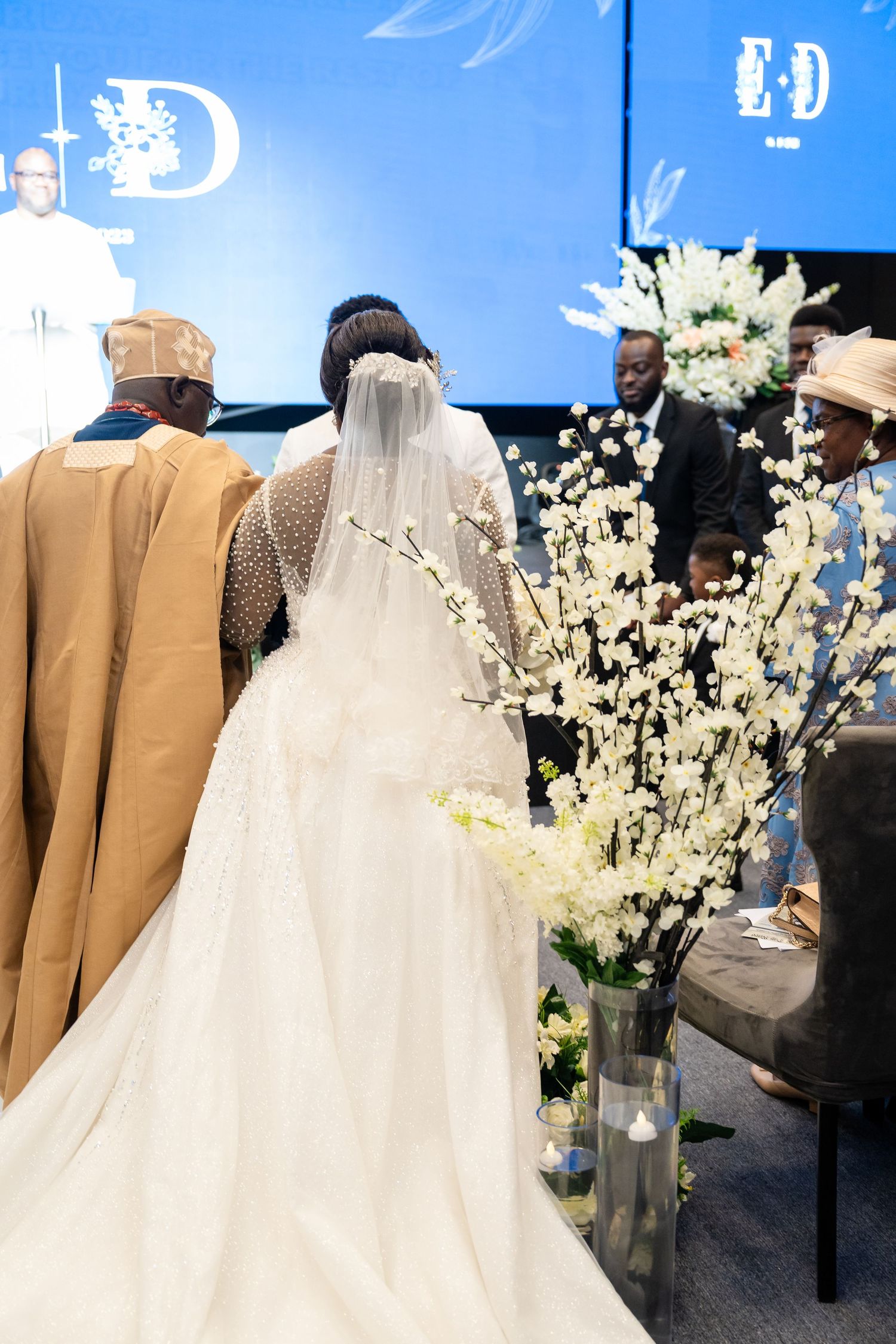 Elegant wedding ceremony setup with tan suits and white floral arrangements against a blue-lit backdrop.