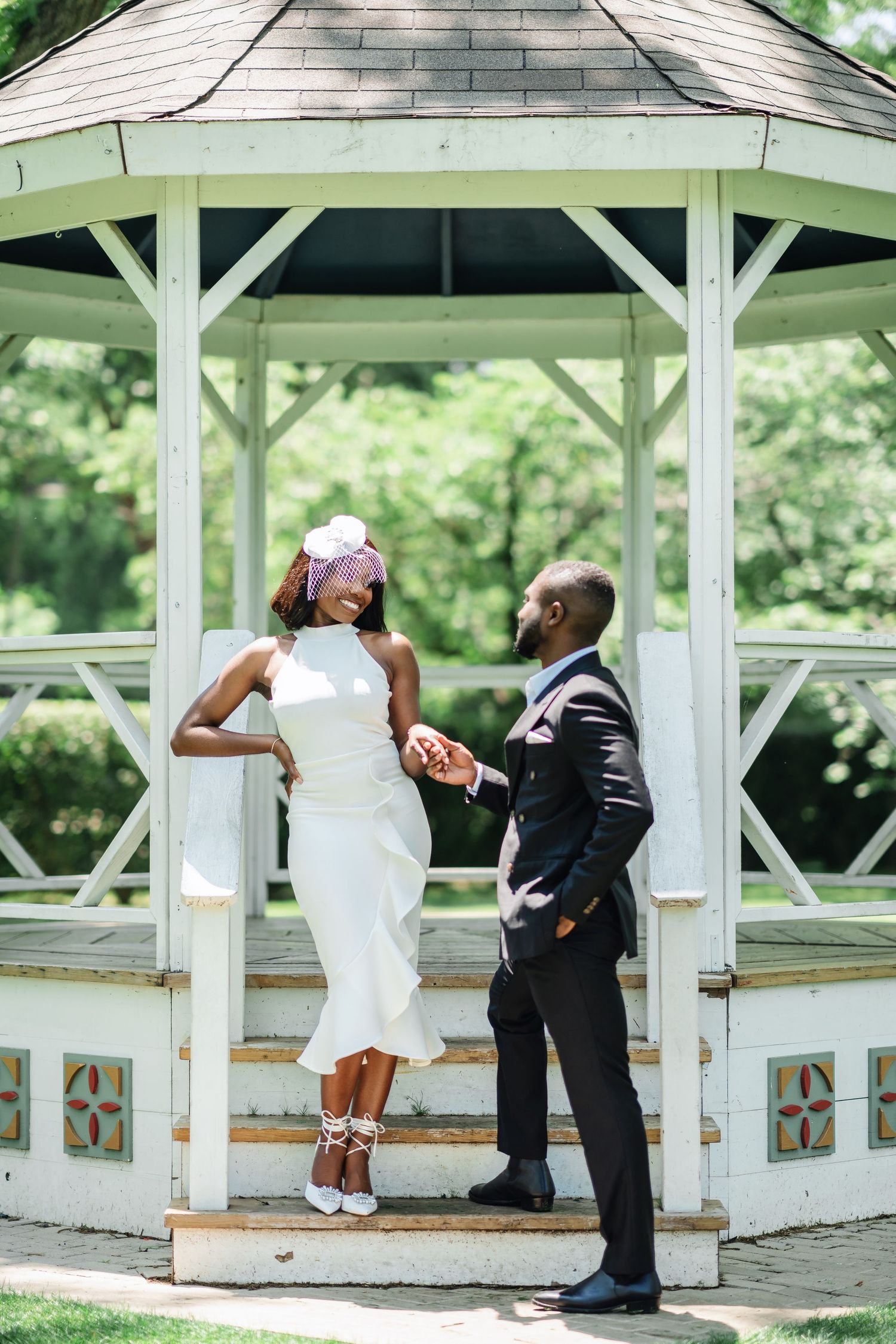 A couple poses playfully in a white gazebo, with the bride in a fitted dress and groom in dark suit.