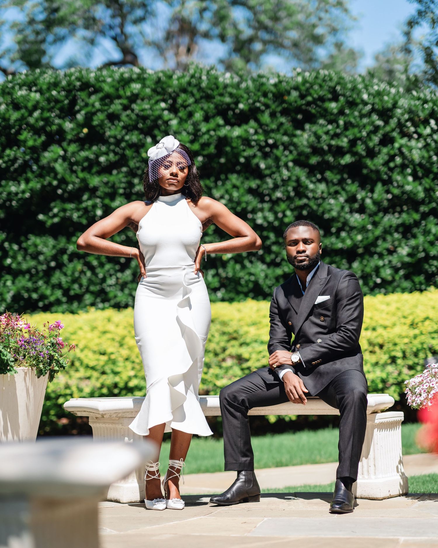 A couple poses playfully during their outdoor wedding photoshoot with manicured gardens and shrubs in the background.