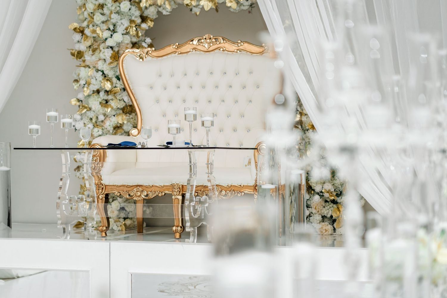 Ornate wedding venue setup with white draping, gold accents, and crystal chandeliers creating a romantic atmosphere.