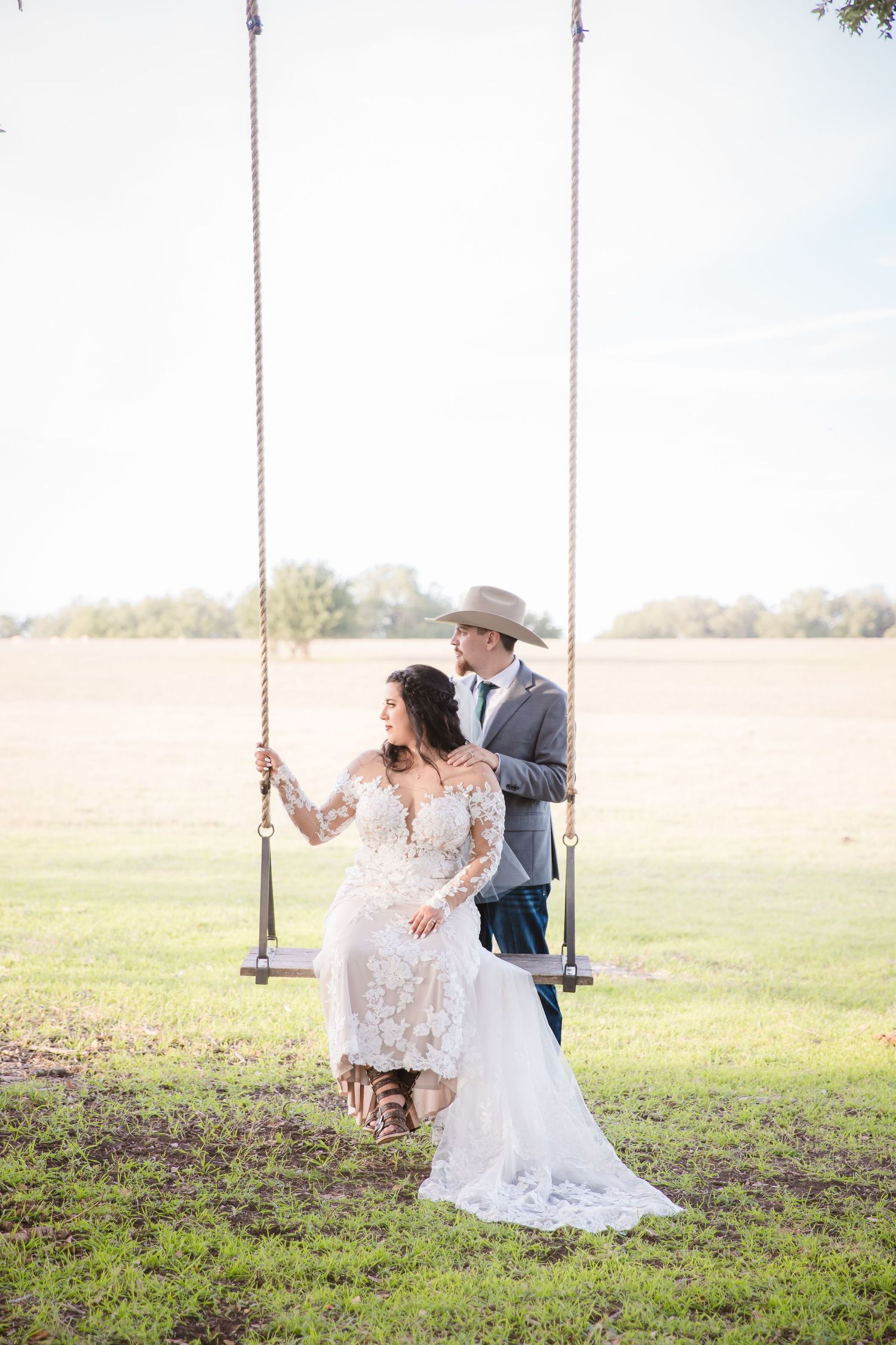 A romantic wedding photo of a couple on a rustic swing in an open field with soft natural lighting.