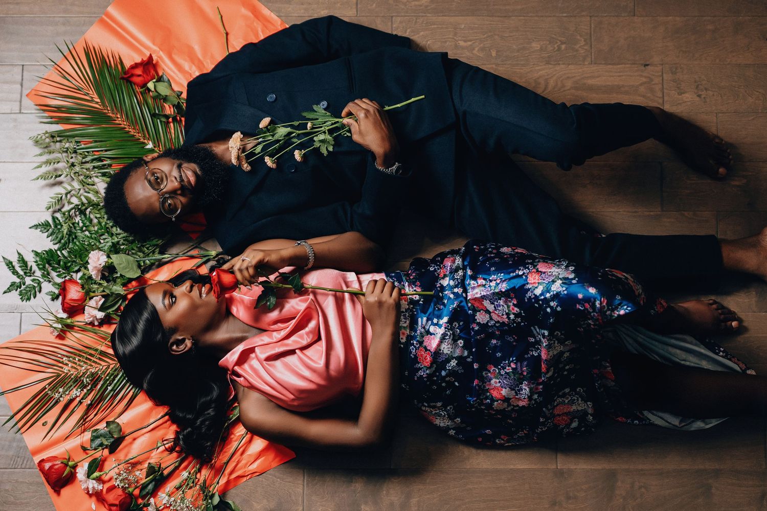 A couple lying on wooden floor surrounded by tropical leaves and orange decor in artistic pose.