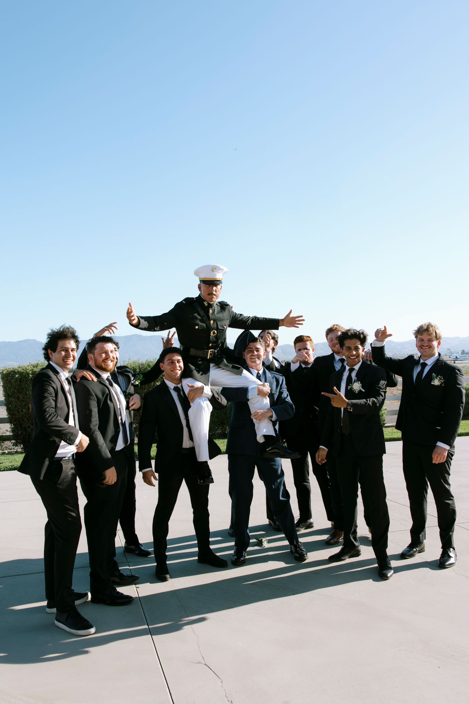 Group of formally dressed people performing a coordinated leaning pose outdoors under blue sky.