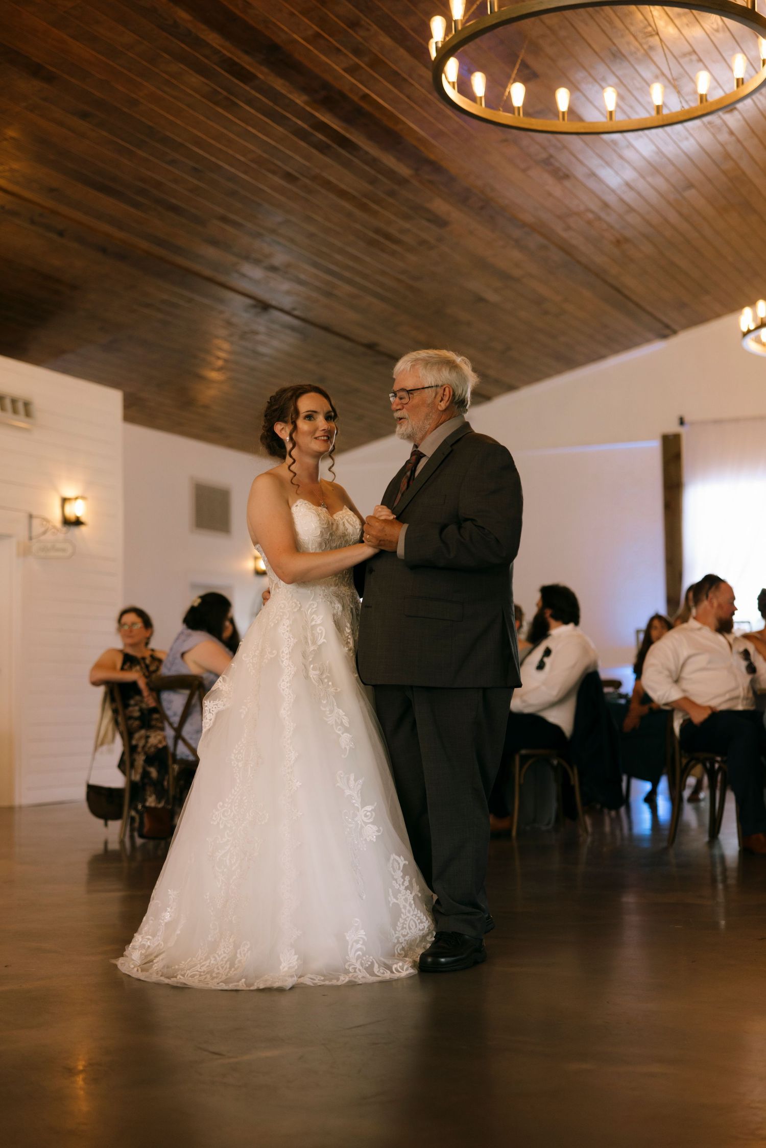 Bride in white lace gown shares father-daughter dance at elegant wedding reception with wood ceiling.