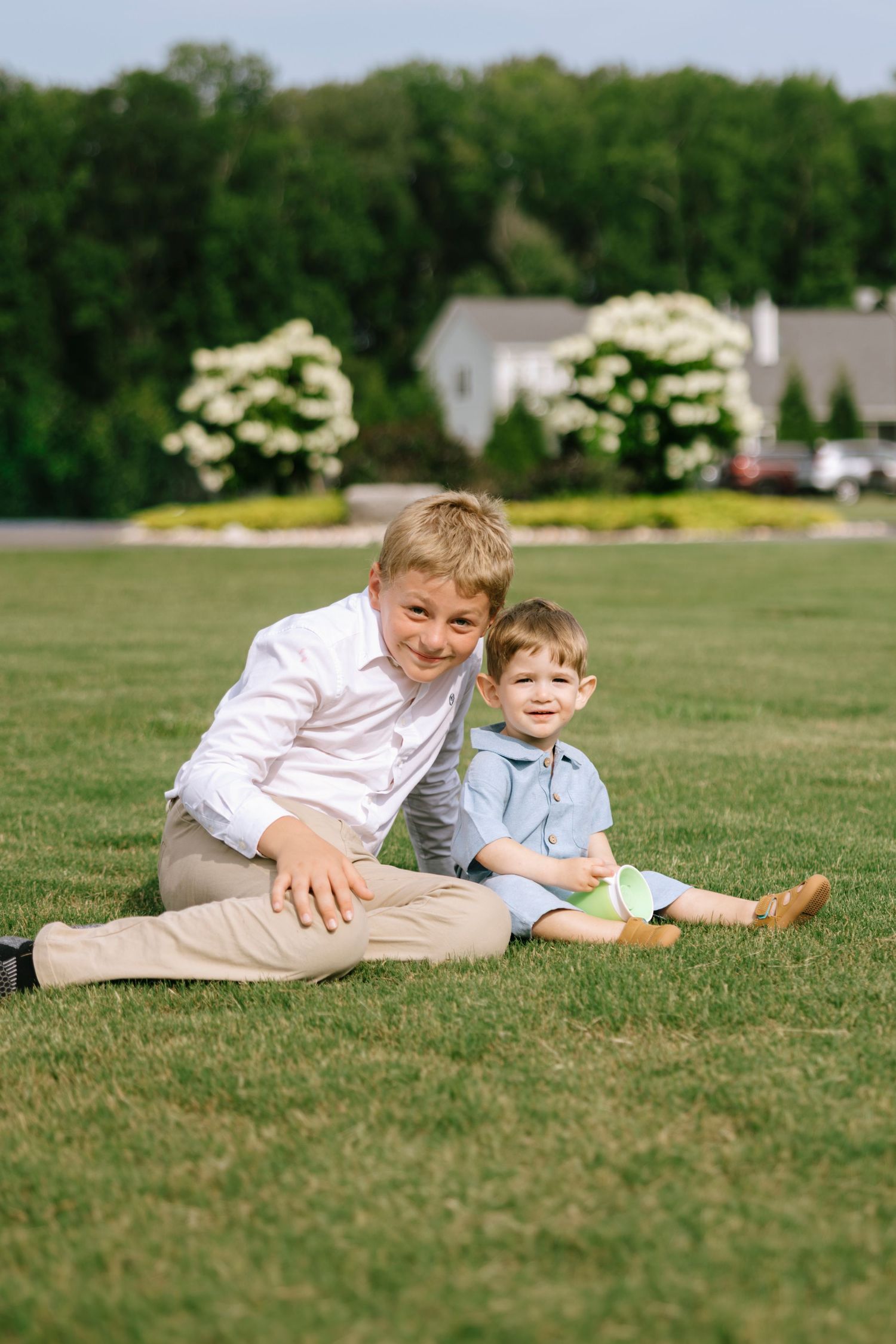 Two boys sitting on green grass outdoors, smiling at camera, with white flowering bushes and trees in background.