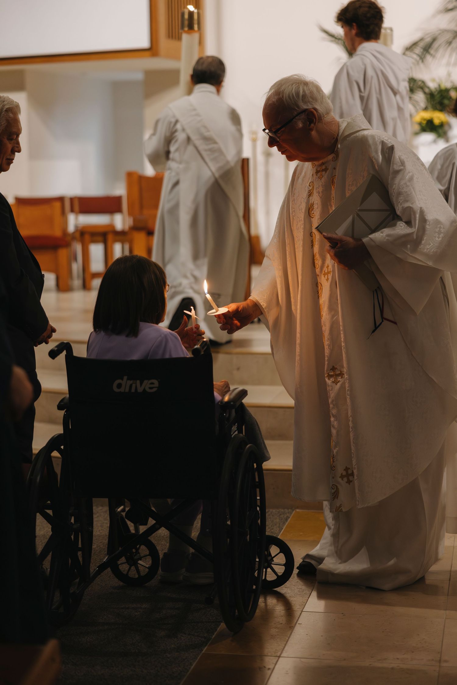 Priest lighting candle for wheelchair-bound parishioner during Catholic Easter Vigil ceremony in church.