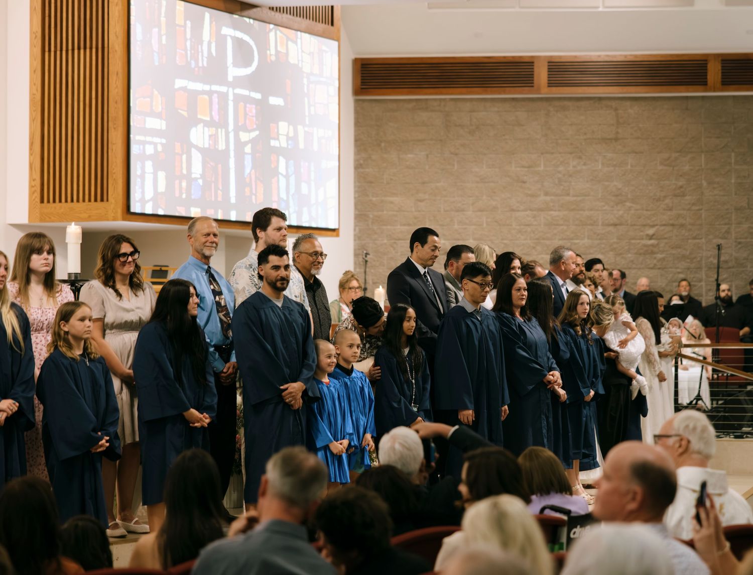 Group of adults and children in blue graduation gowns standing at the front of a church during a religious ceremony.