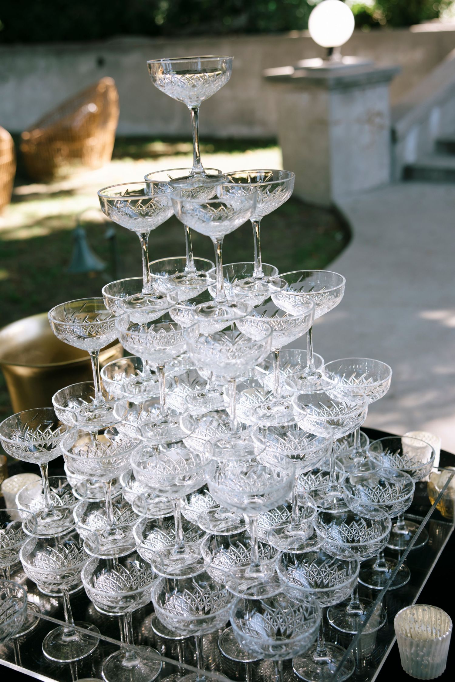 Crystal champagne coupes are stacked in elegant pyramid formation at an outdoor event.