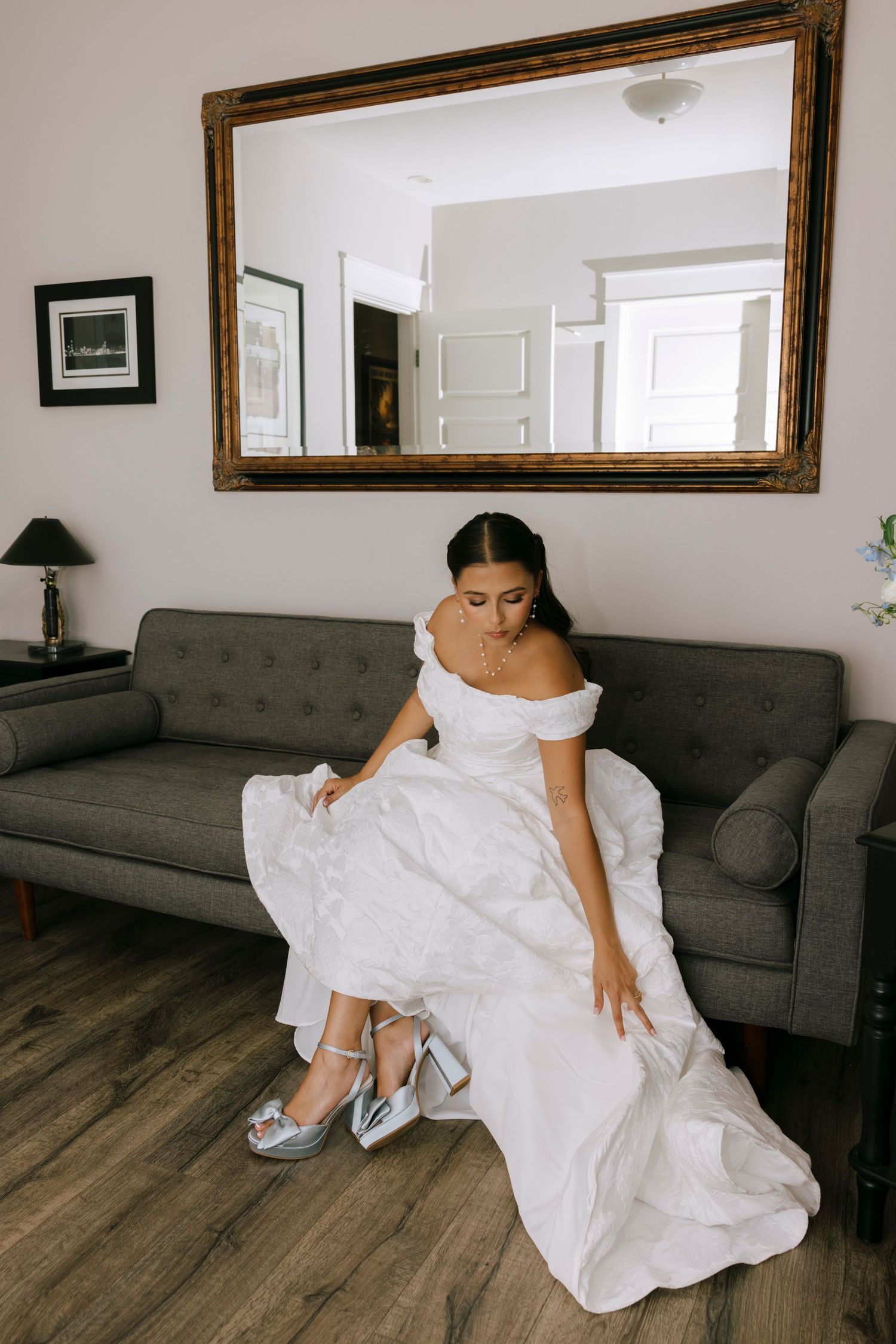 A bride in a white gown rests on a grey couch while showing off blue shoes.