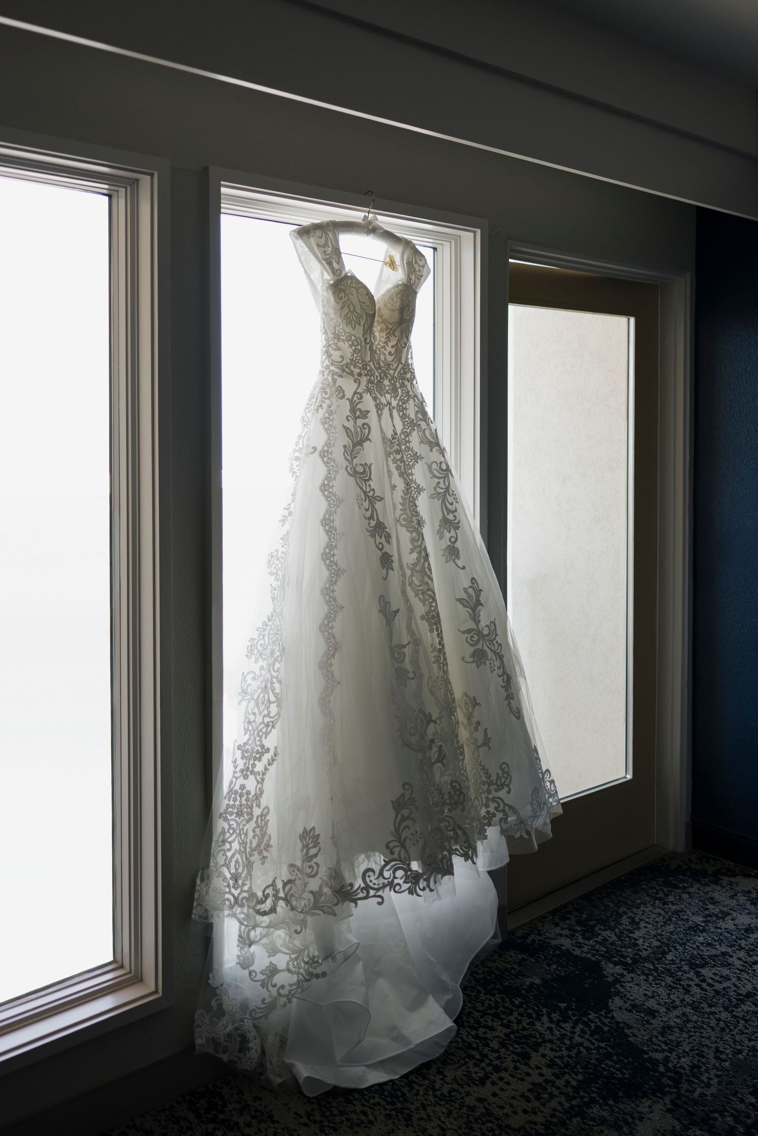 Elegant lace wedding gown with floral embroidery hanging on a window hanger in a softly lit bridal suite.