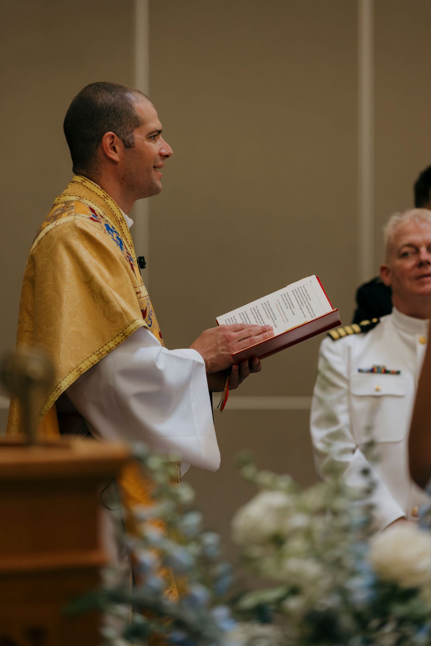 Catholic priest in gold vestments holds a book during a religious ceremony beside a naval officer in white uniform.