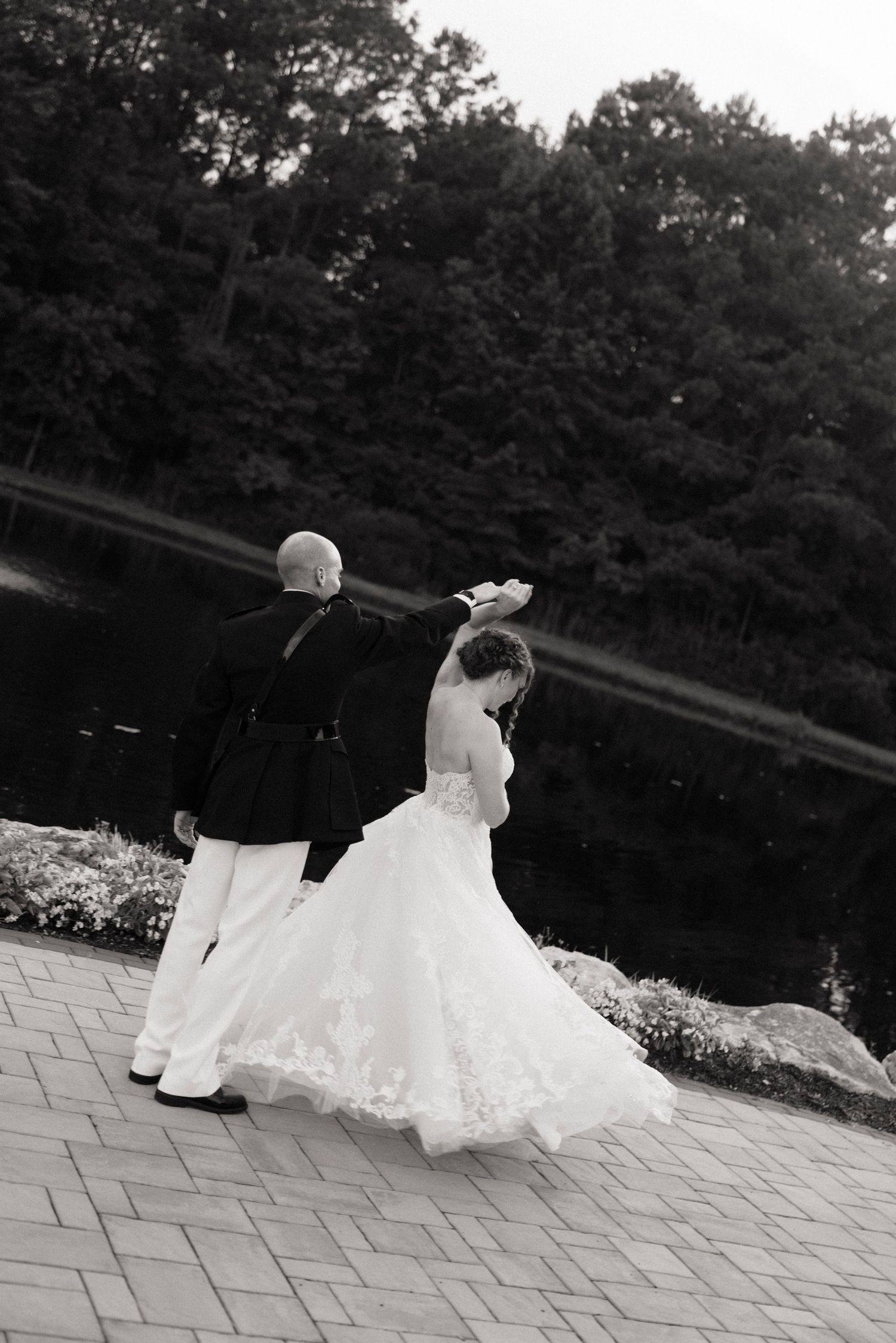 Black and white photo of a bride and groom dancing outdoors near a pond, surrounded by lush trees.