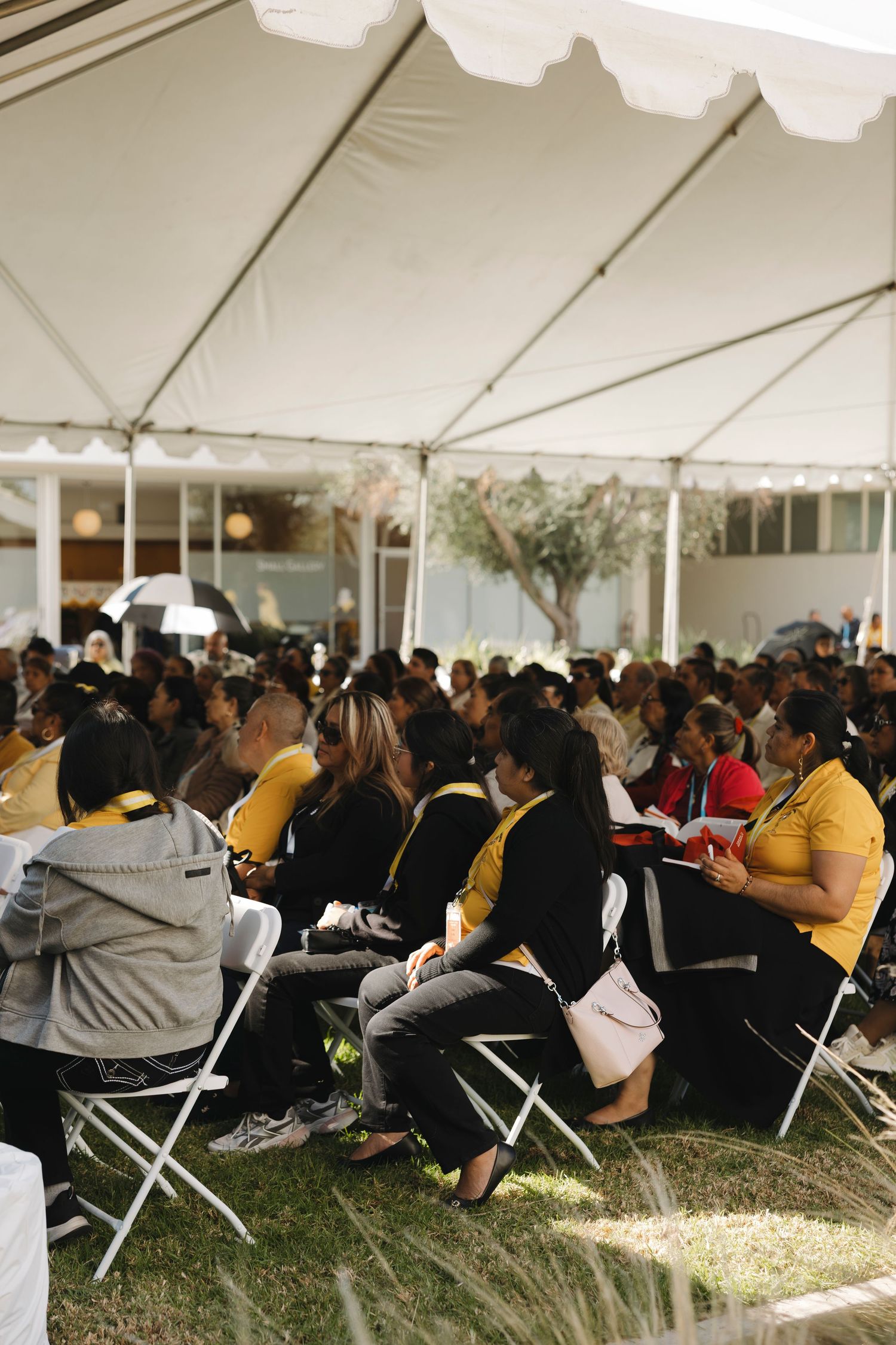 Large crowd seated under white tent during outdoor conference or seminar.