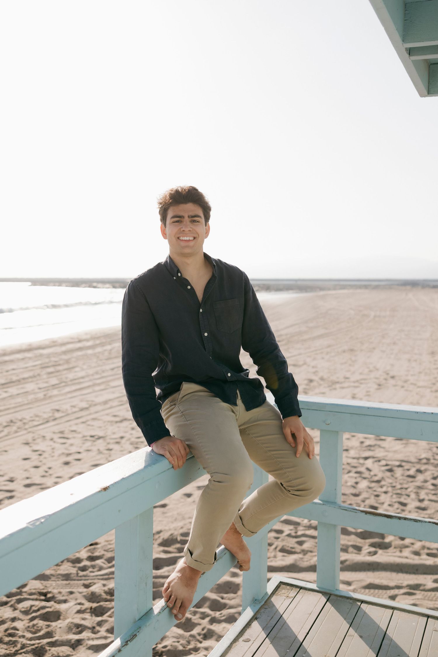 Young man in black shirt and khaki pants sitting on a light blue beach railing, smiling at camera.