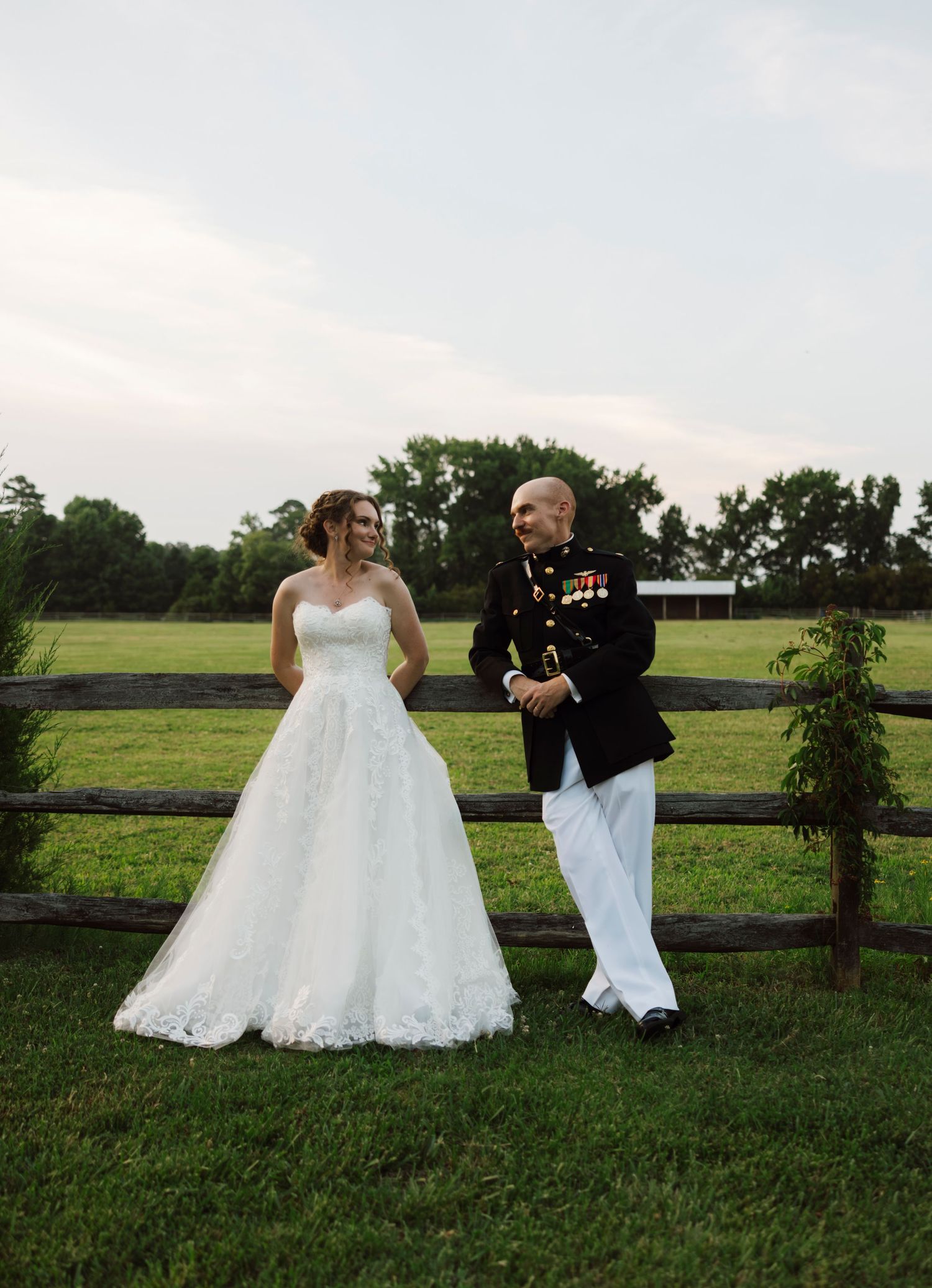 A bride in a white lace gown and a groom in Marine dress uniform stand by a wooden fence on a green farm.