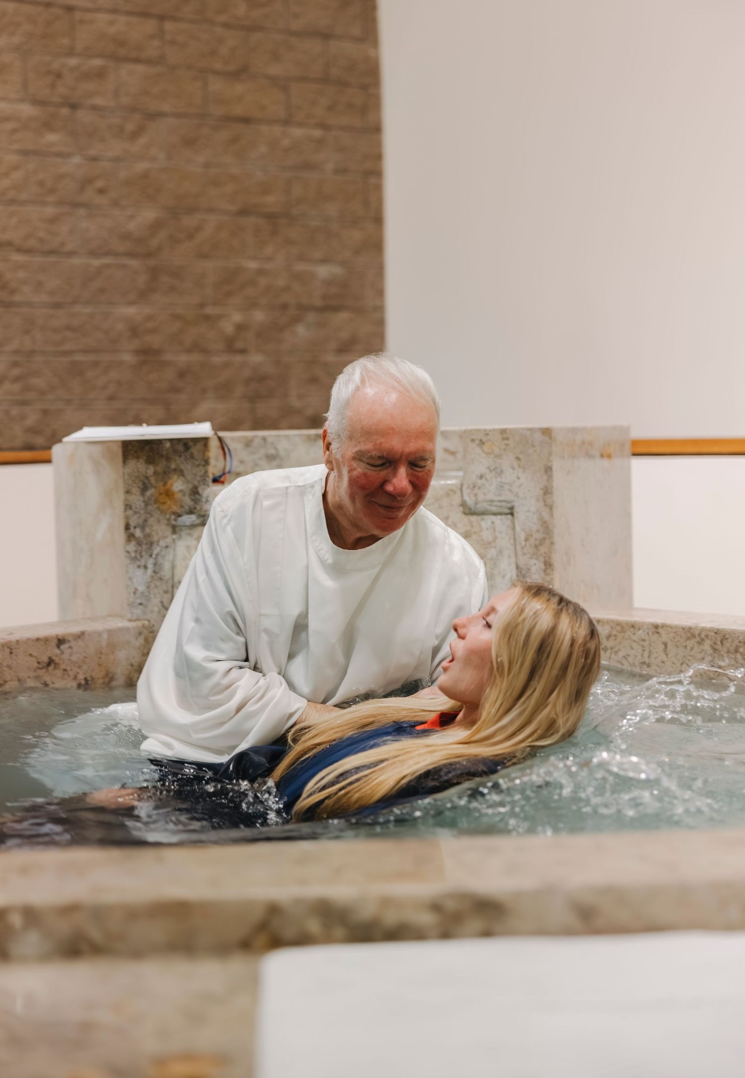Elderly priest performing full immersion baptism with a young woman in a church baptismal pool.